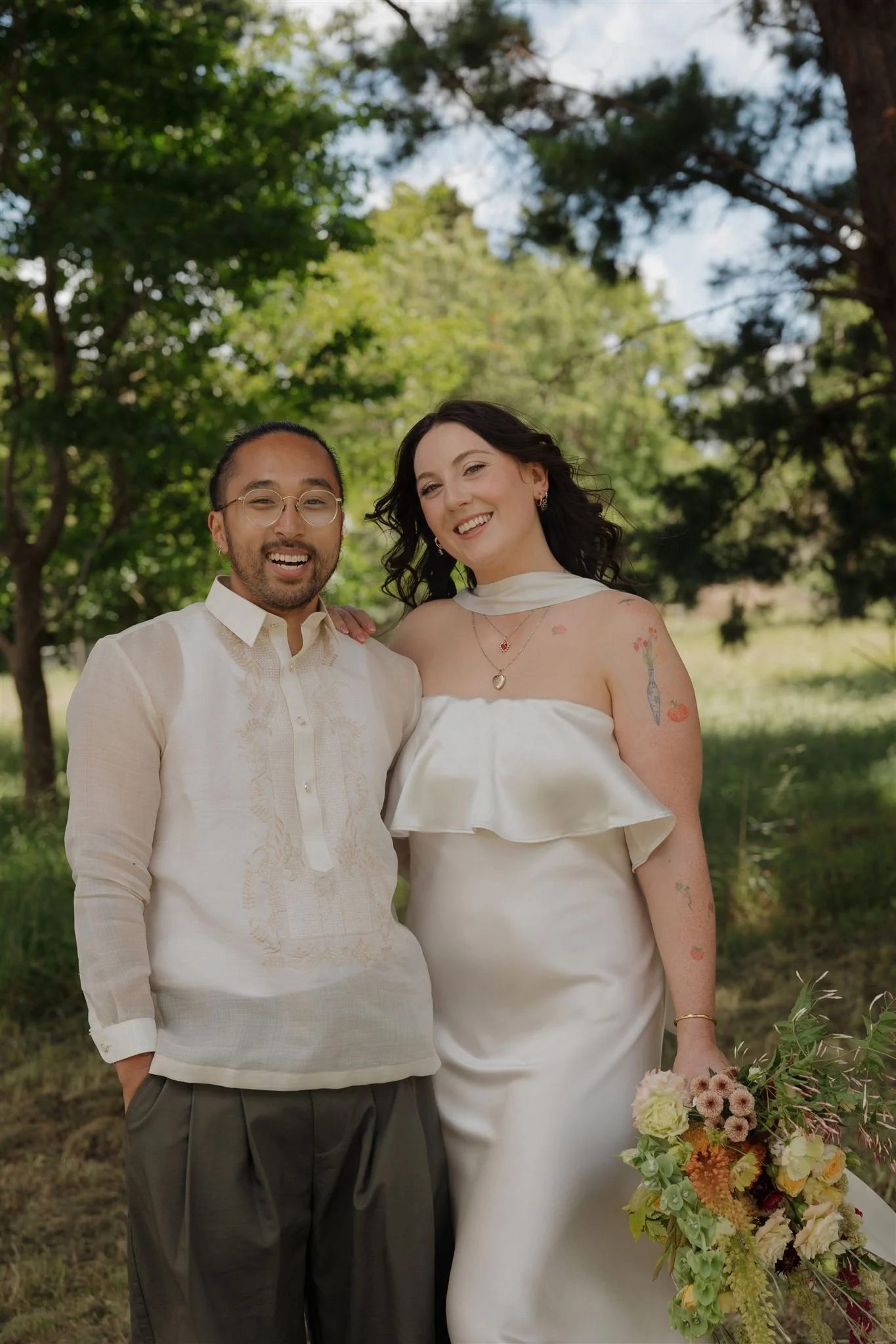A smiling couple standing outdoors among trees, with the woman holding a large bouquet of colorful flowers. Bride and groom. Wellington makeup artist lucy loo did an amazing soft glam on the gorgeous bride.