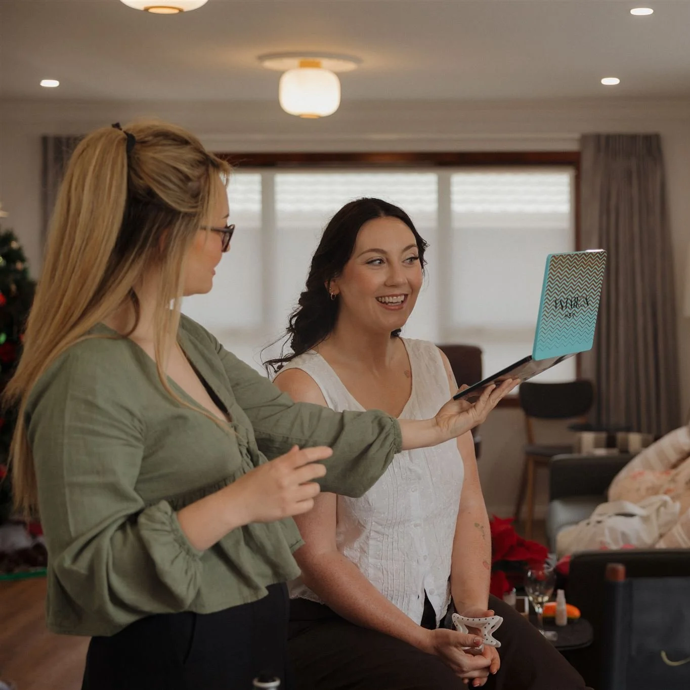 Two women sharing a joyful moment indoors. The woman on the left with long blonde hair, glasses, and a green top is holding a small, striped gift box. The woman on the right with dark hair, using a white sleeveless top, is smiling, holding the gift, and looking at the woman on the left. In the background, there are windows with blinds, curtains, a Christmas tree, and some furniture.