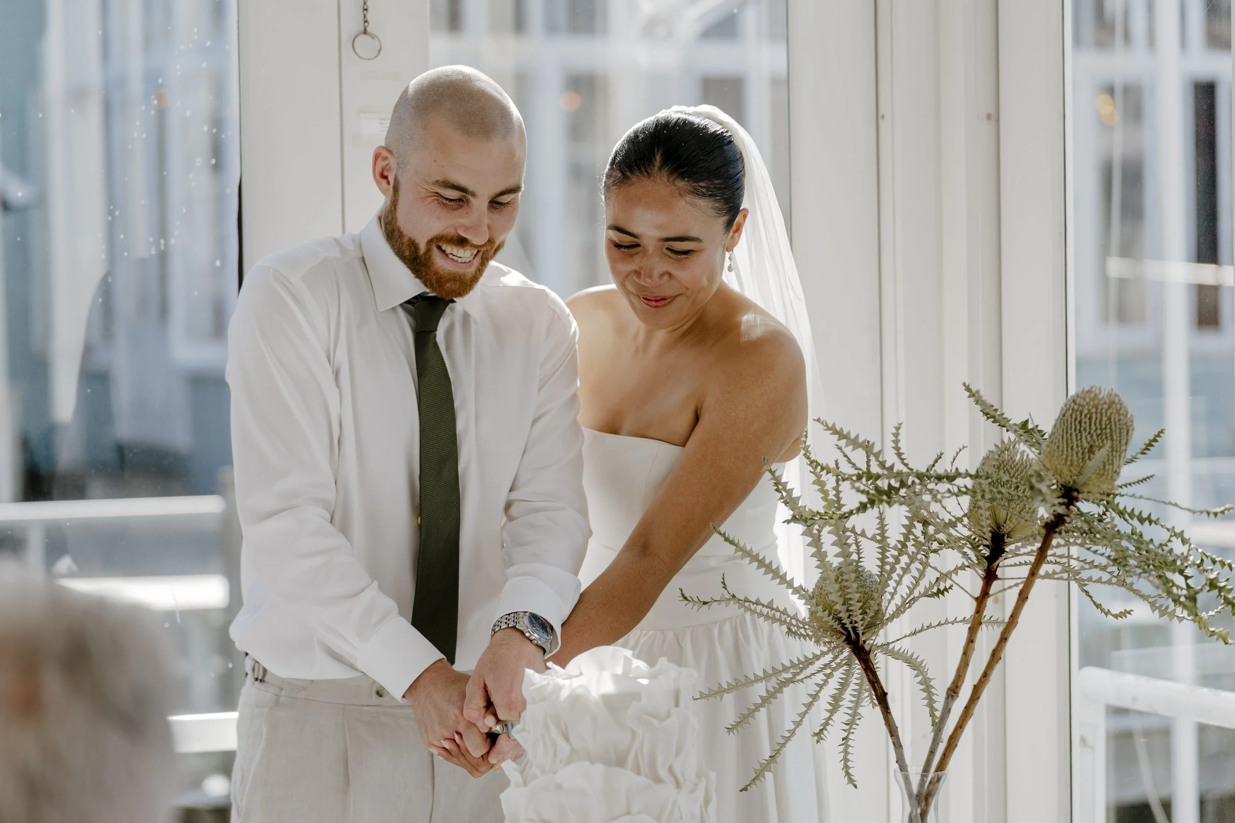 A bride and groom cutting a wedding cake together indoors near a large window, with a floral arrangement in the foreground. Natural makeup look. soft glam, natural glam. Makeup by lucy loo
