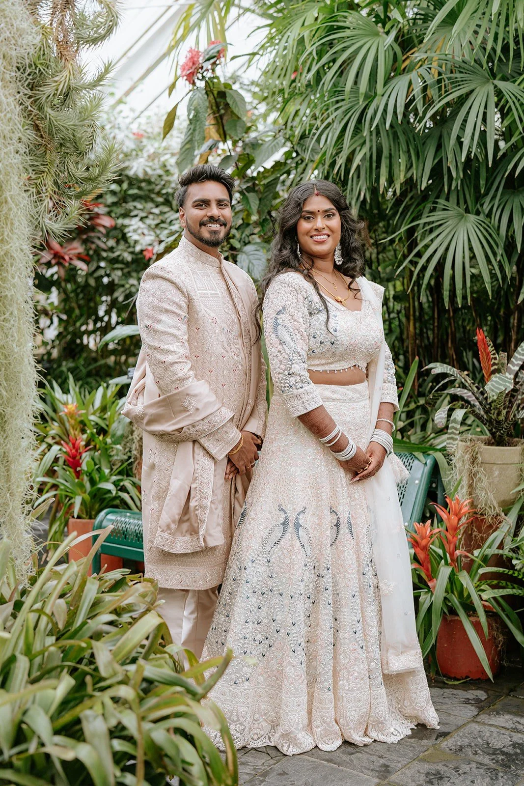 A couple dressed in traditional Indian attire standing in a lush green greenhouse or garden surrounded by various tropical plants and flowers. Indian wedding. Indian bridal makeup. Full glam makeup look. Contouring, flawless application wellington nz