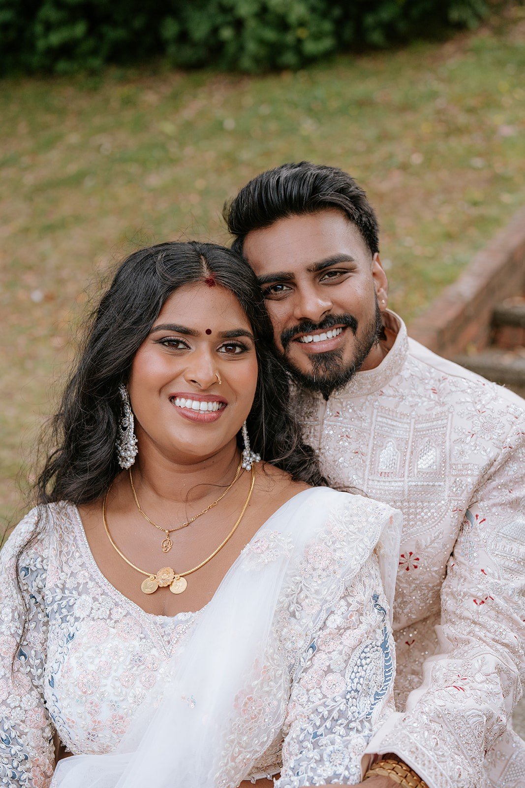 A smiling Indian couple dressed in traditional attire, outdoors with greenery background, taking a close-up selfie. Indian wedding. Indian bridal makeup. Flawless, makeup by wellington makeup artist. Professional makeup for women of colour