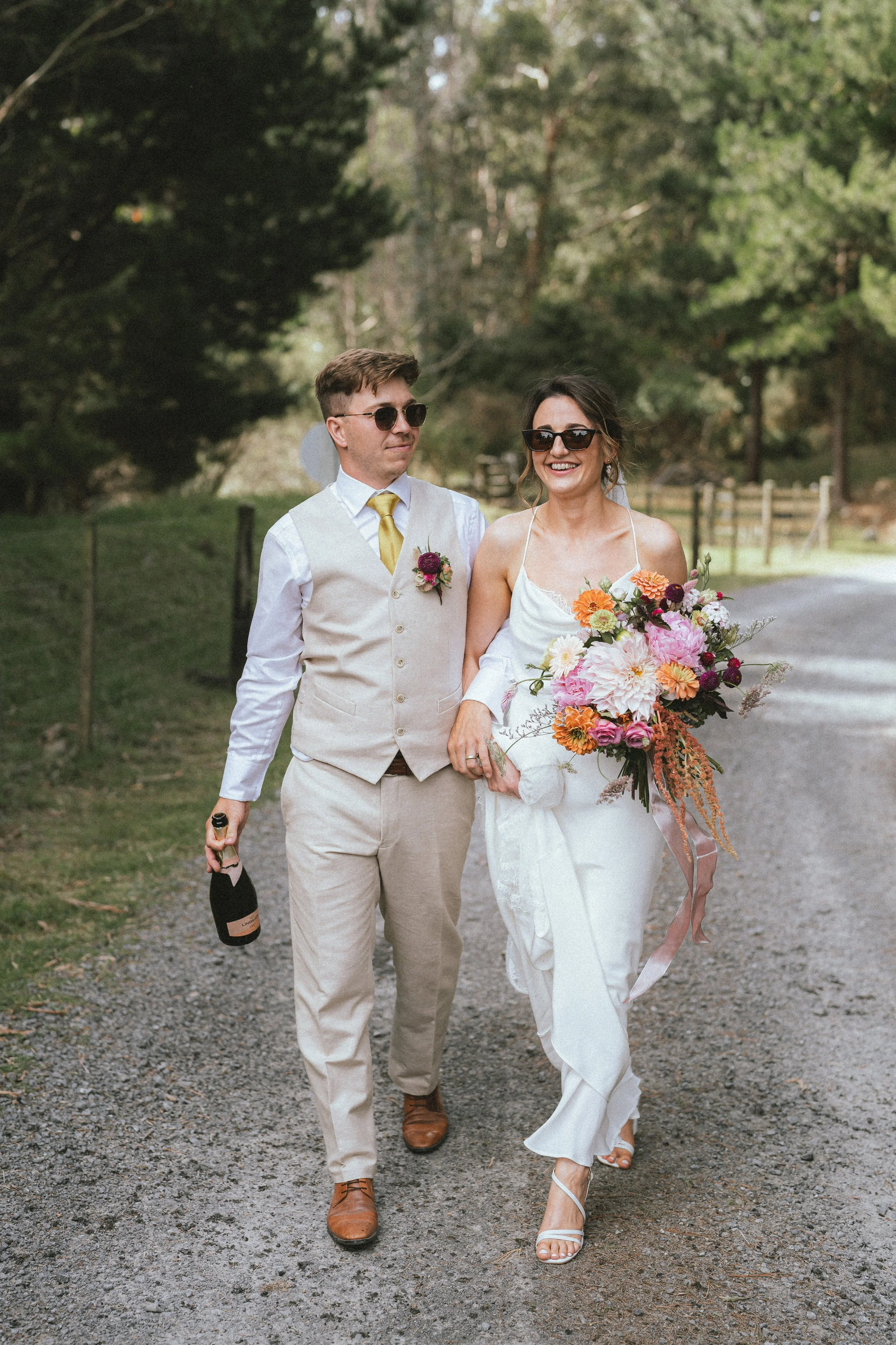A bride and groom walking outdoors on a gravel path, surrounded by greenery and trees in daylight. The bride is holding a large, colorful bouquet and wearing sunglasses, while the groom is holding a bottle of champagne and wearing sunglasses.