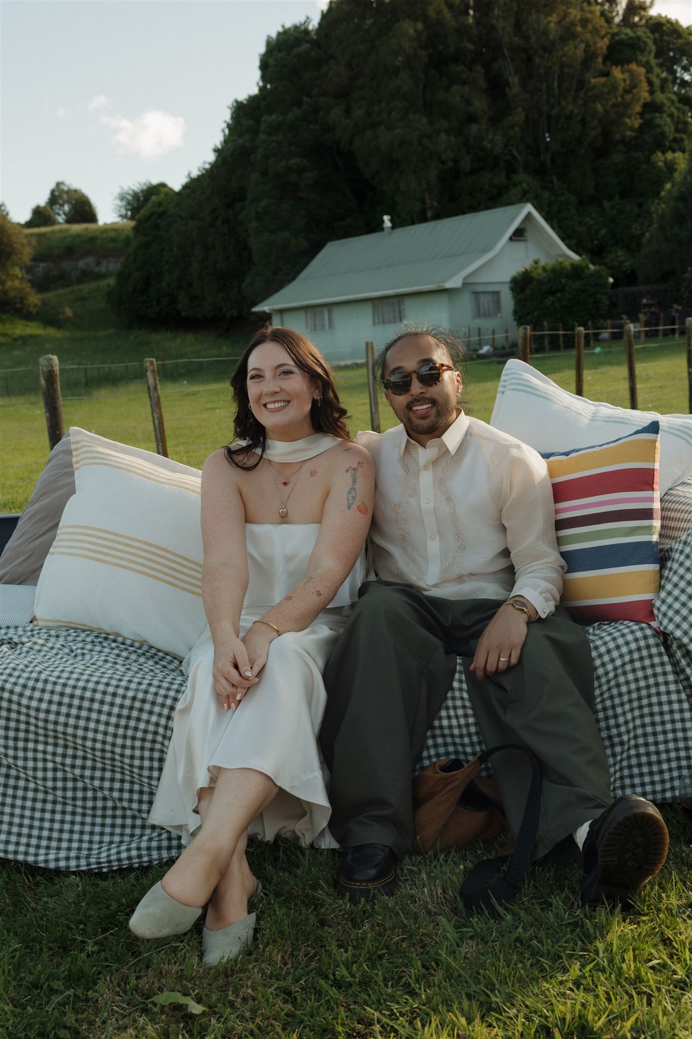 A smiling woman and man sitting together outdoors on cushions, with a house and lush green trees in the background. bride and groom