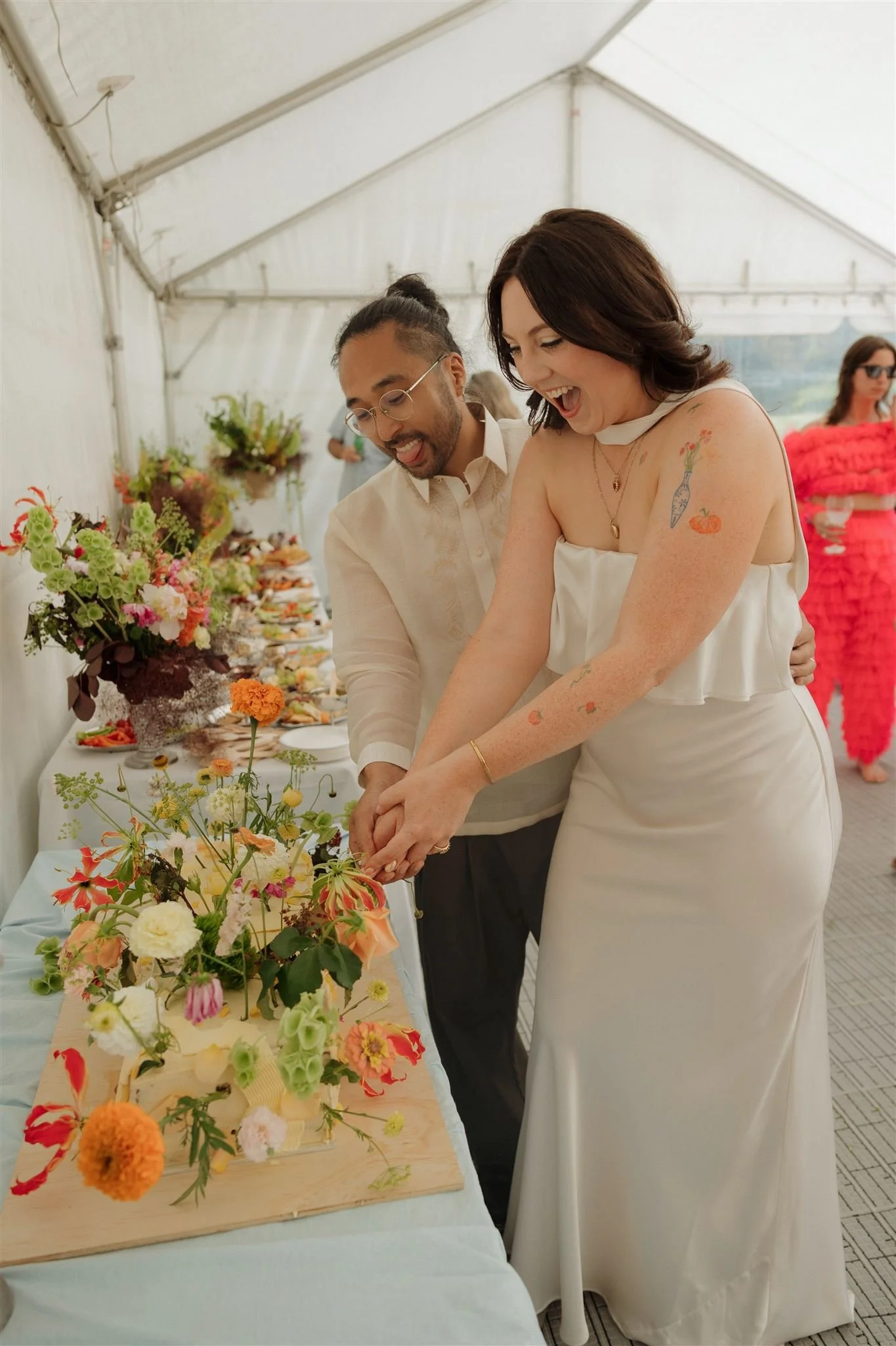 A woman in a white dress with colorful tattoos on her arm is smiling and cutting a cake at a celebration, with a man beside her. The table has various colorful floral arrangements and desserts, set in a bright outdoor tent. Bride and groom