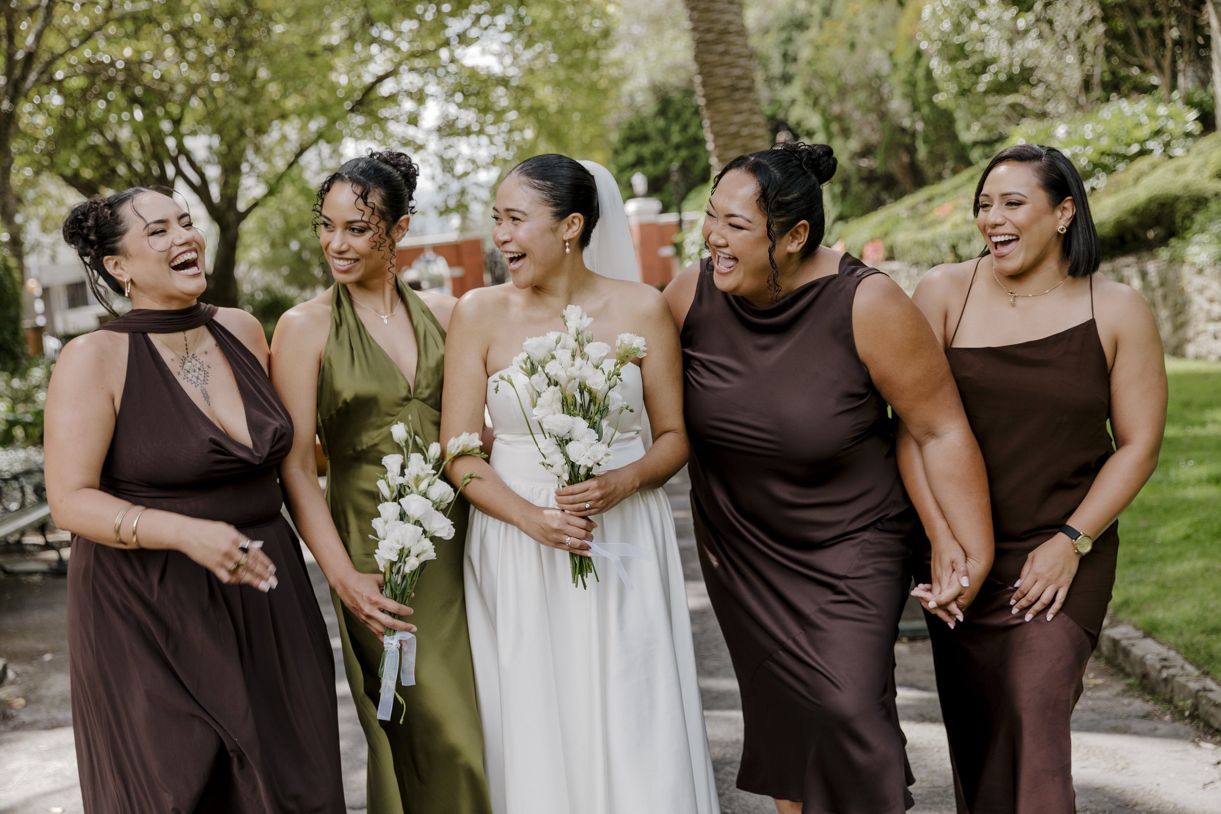 Group of five women, including a bride holding white flowers, smiling and laughing outdoors on a sunny day. Bride with bridesmaids. Makeup and hair done. Wedding makeup wellington makeup artist