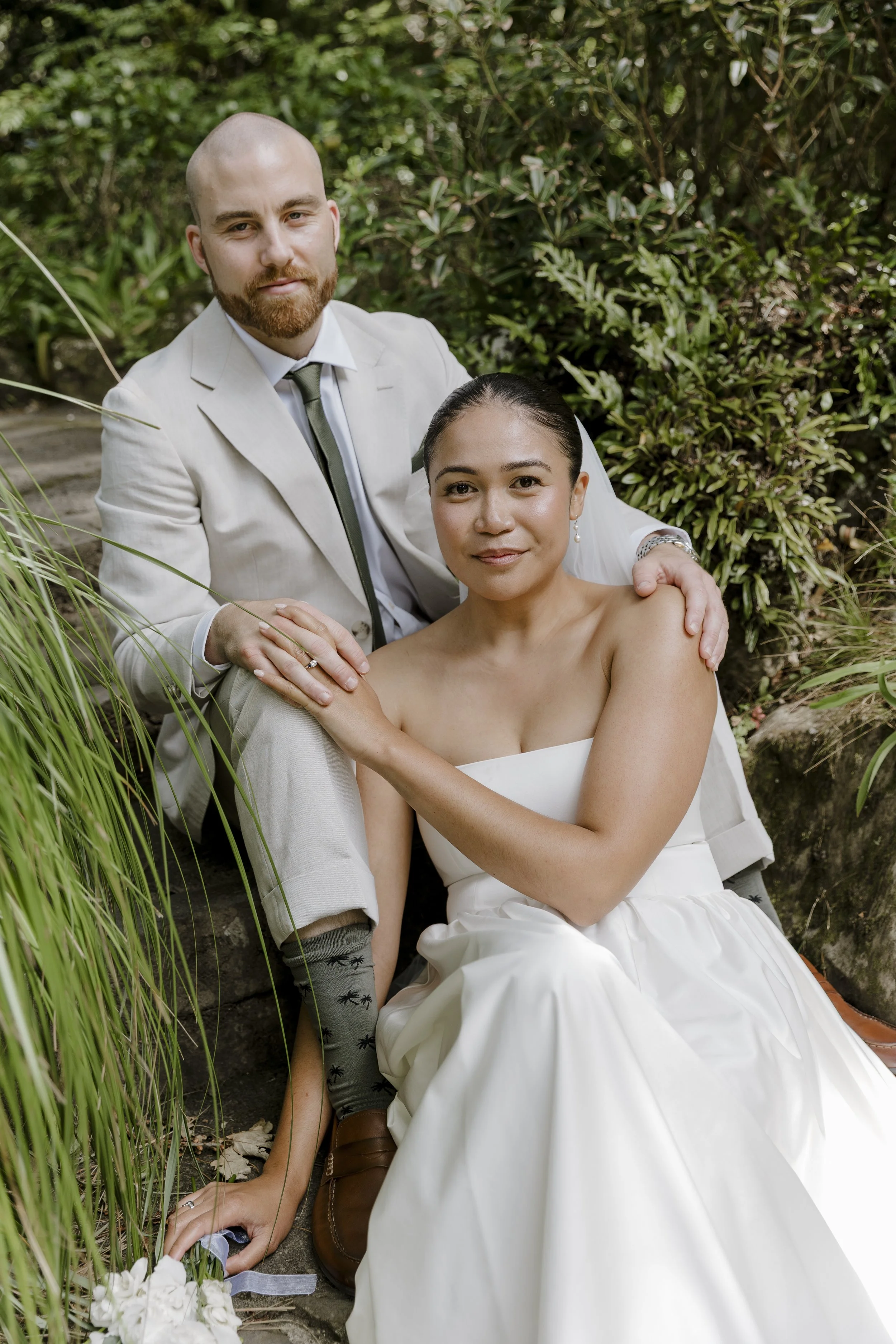 A man and a woman sitting outdoors in a garden, posing for a wedding photo. The man is wearing a white suit with a tie and has a beard, while the woman is dressed in a white strapless wedding gown and has her hair pulled back. They are surrounded by 