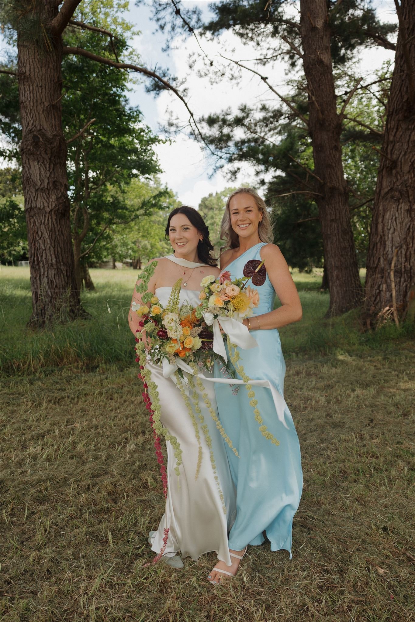 Two women in dresses are standing together outdoors on grass, holding a large bouquet of flowers. They are smiling, with trees and a partly cloudy sky in the background. Camera ready makeup. Flawless soft glam, natural glam. 