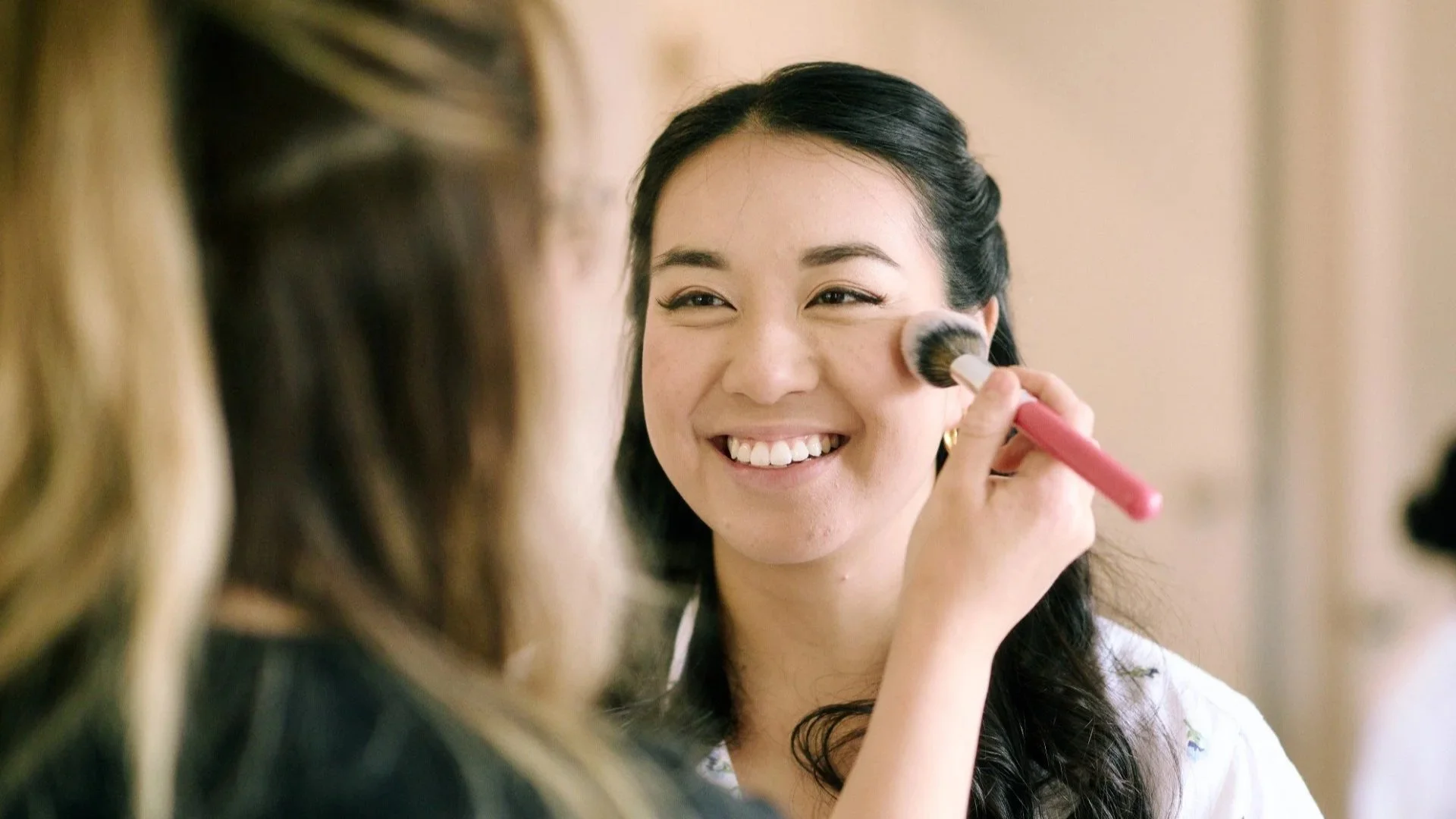 A woman smiling as another woman applies blush to her cheek with a makeup brush.