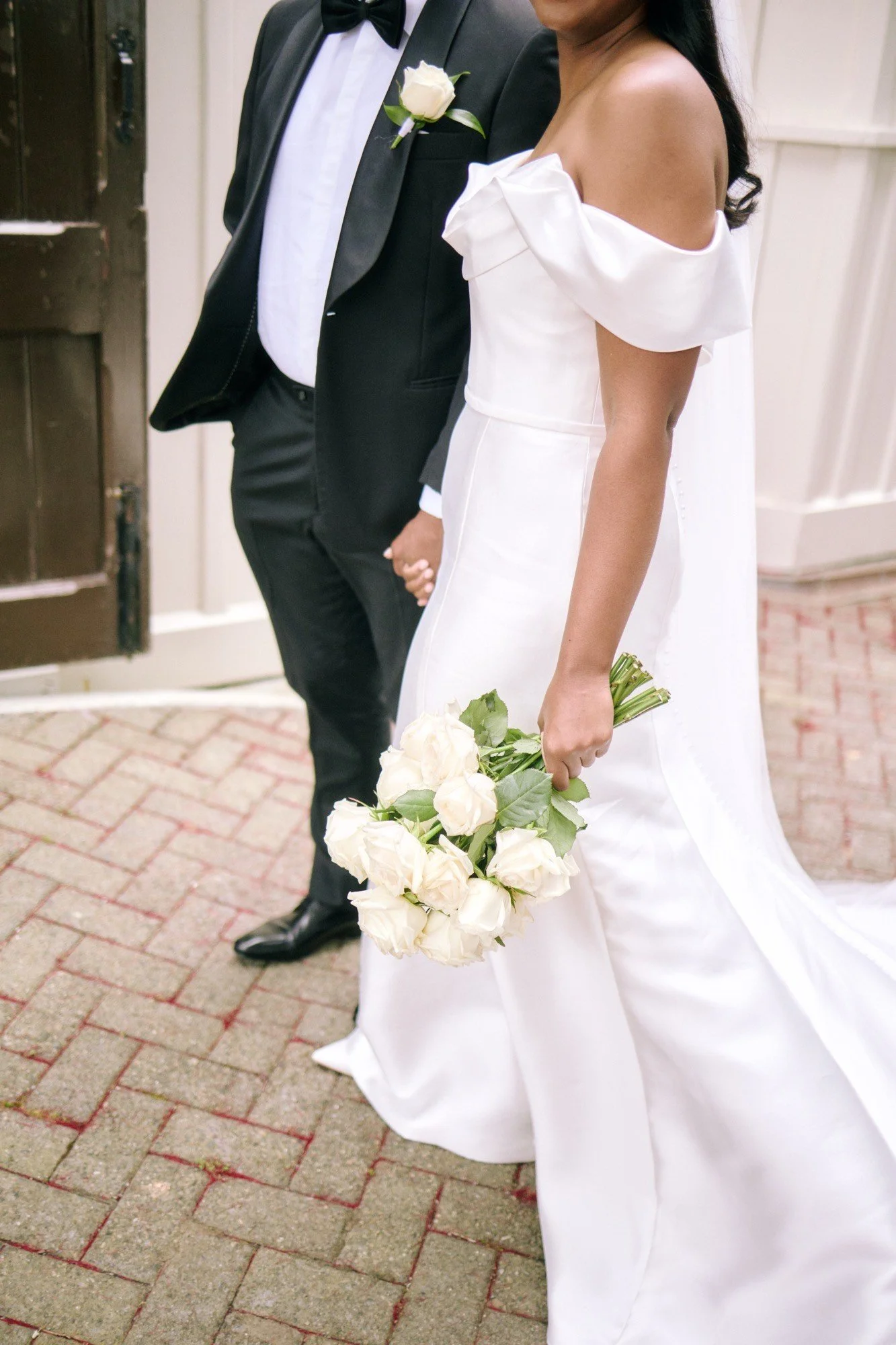 A bride and groom holding hands, with the bride holding a bouquet of white roses. The bride is wearing a white wedding dress with off-the-shoulder sleeves, and the groom is in a black tuxedo with a white shirt and black bow tie. They are standing on a brick walkway, partially visible from the waist down.