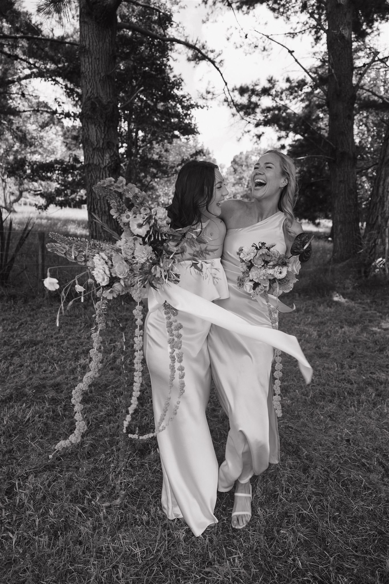 Two women, dressed in wedding gowns, are standing close together outdoors surrounded by trees. They are laughing and holding bouquets of flowers, appearing joyful and happy. Beautiful bridal makeup look. Bridal party. Natural makeup.