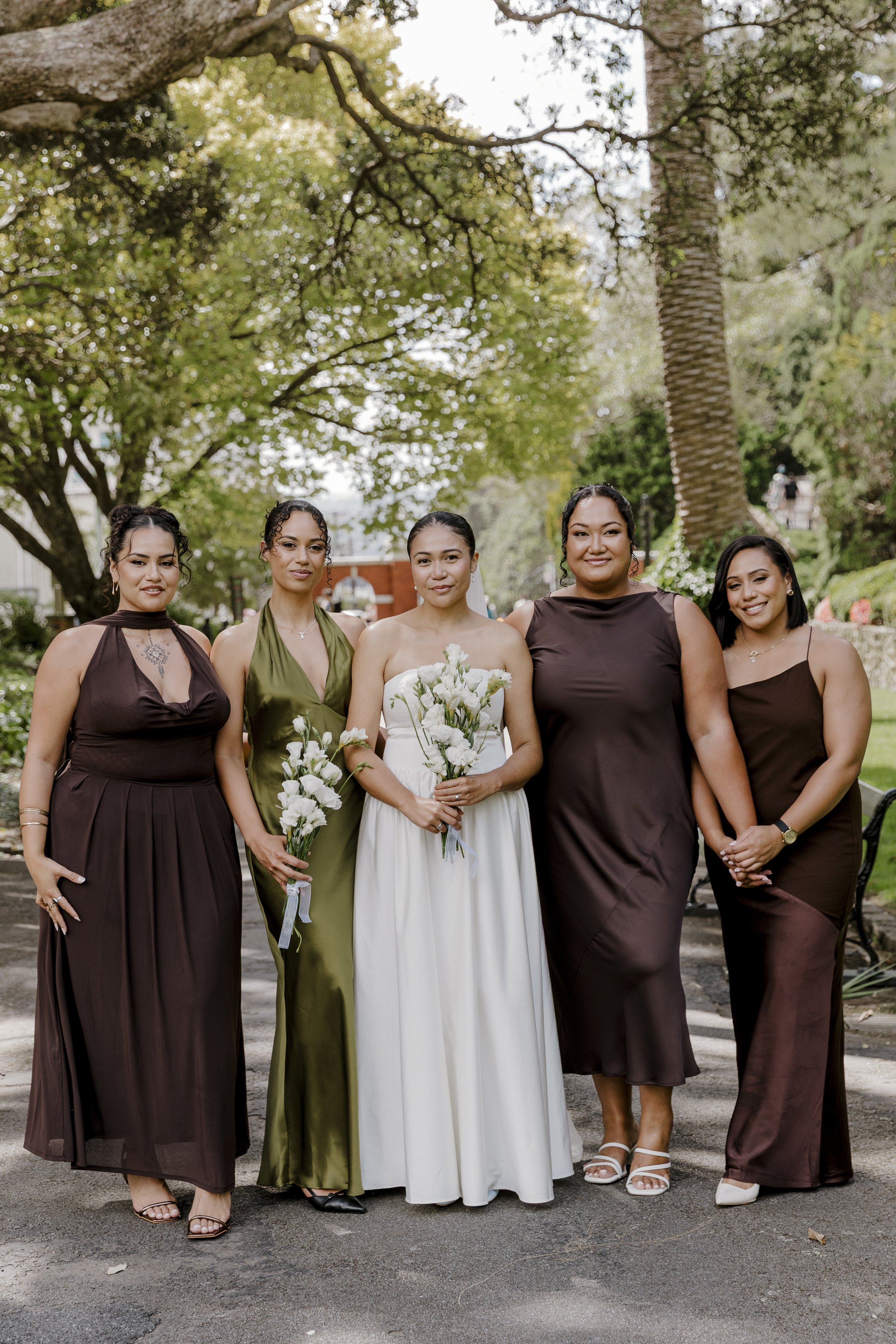 Five women dressed in formal attire standing outdoors under trees, one woman in the center is holding a bouquet and is likely a bride, with the others possibly bridesmaids. Bride with bridal party. Flawless makeup by lucy loo wellington makeup artist