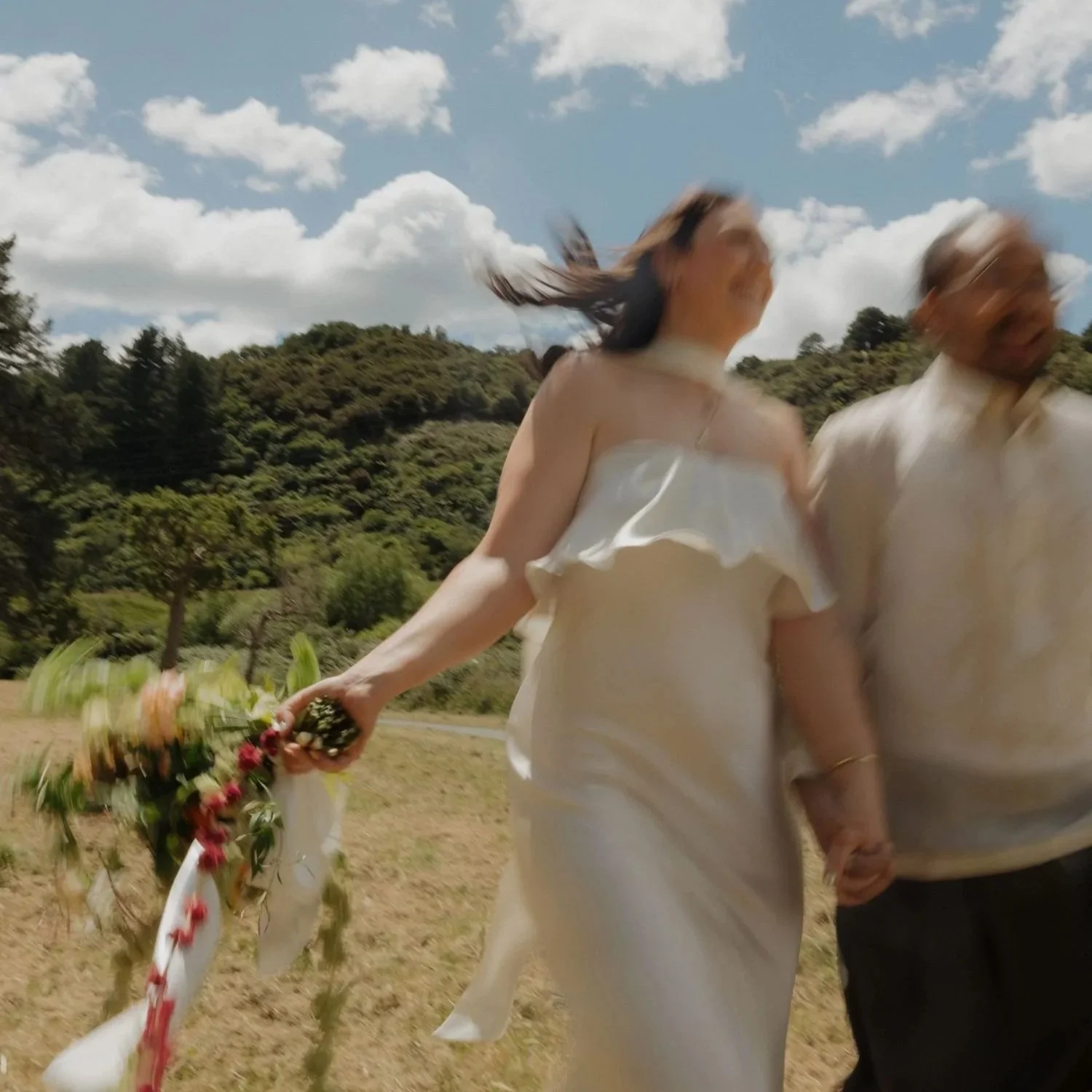Two people holding hands outdoors with a scenic background of trees, hills, and a partly cloudy sky, with the woman holding a bouquet of flowers. Bride and groom