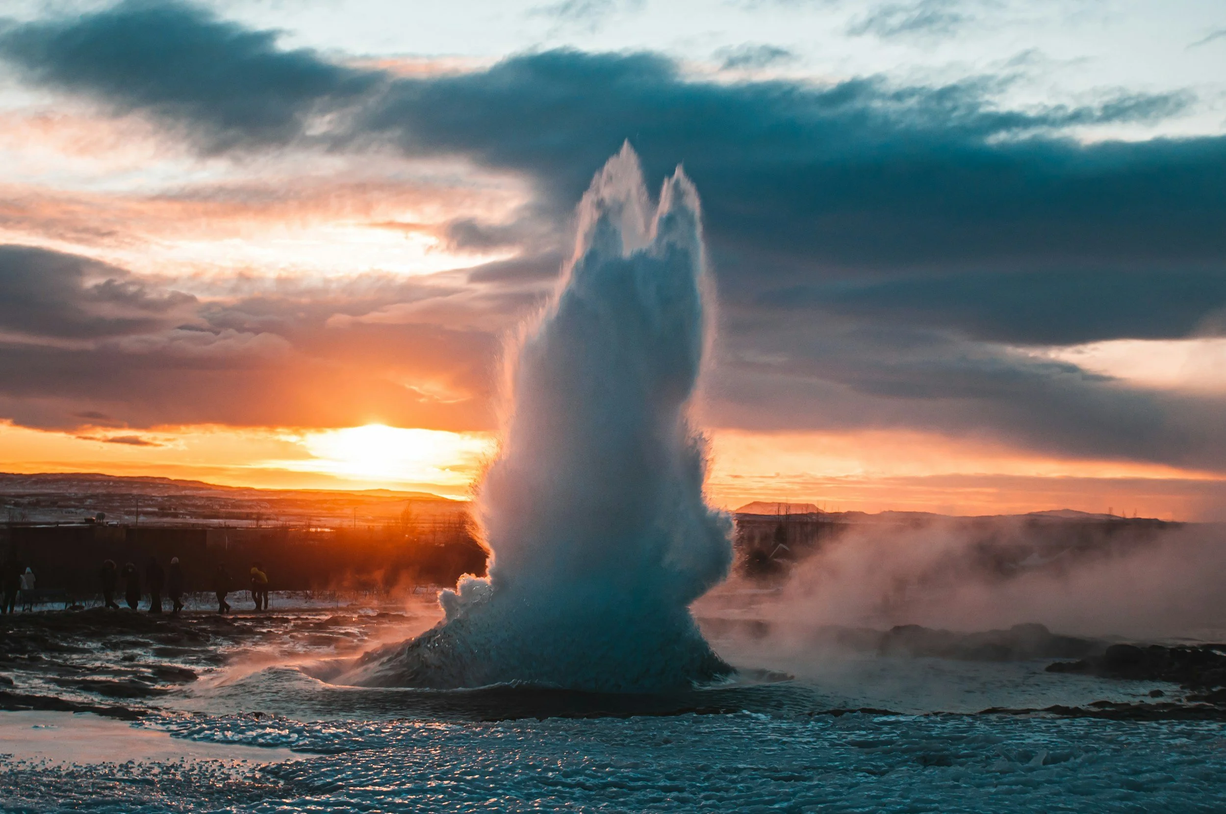 A large fountain jet of water shooting high into the sky against a sunset, with dark clouds and orange light in the background, with people observing from the shoreline.