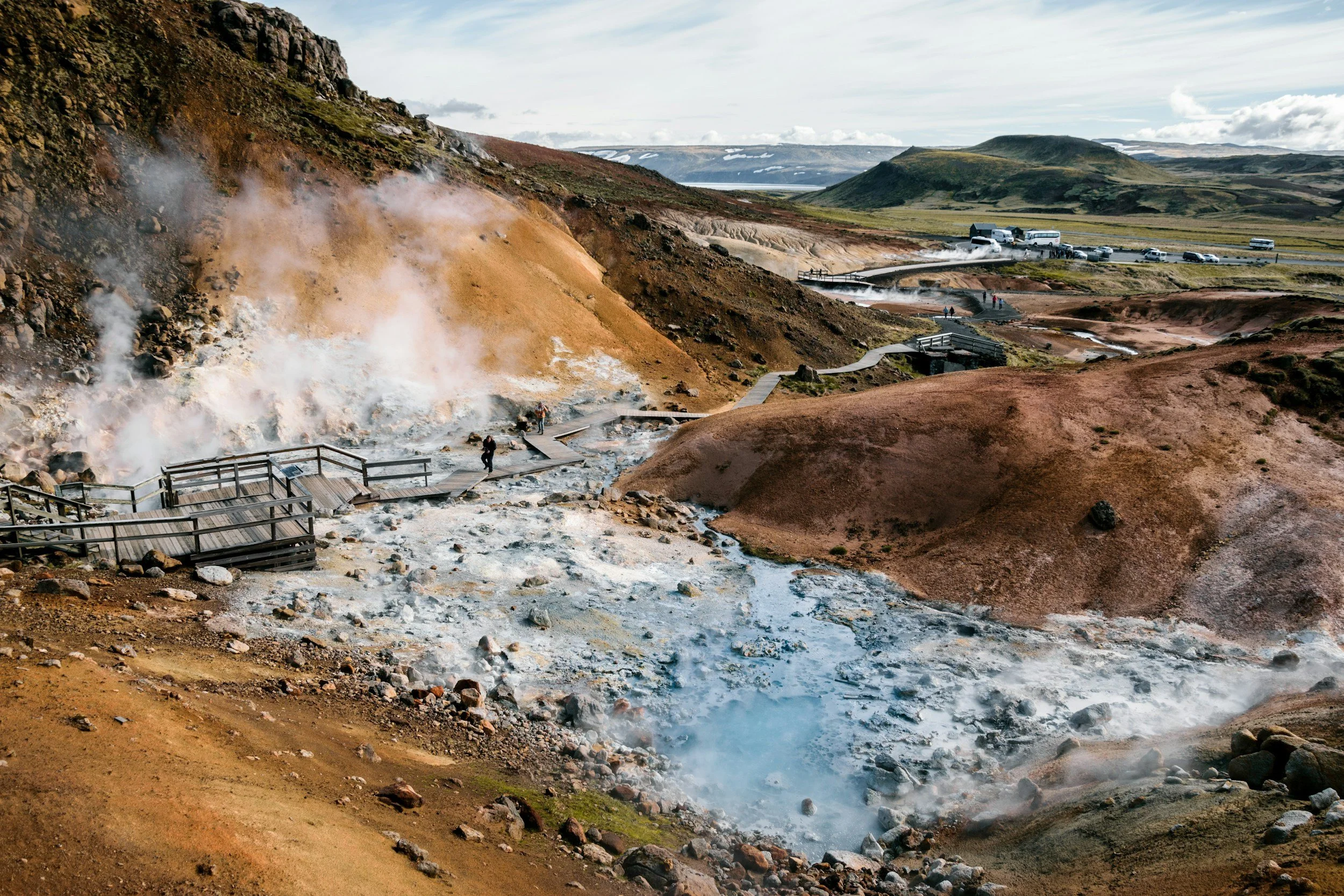 A geothermal area with steaming vents and colorful mineral deposits, surrounded by a mountainous landscape with a viewing walkway and parked vehicles in the background.