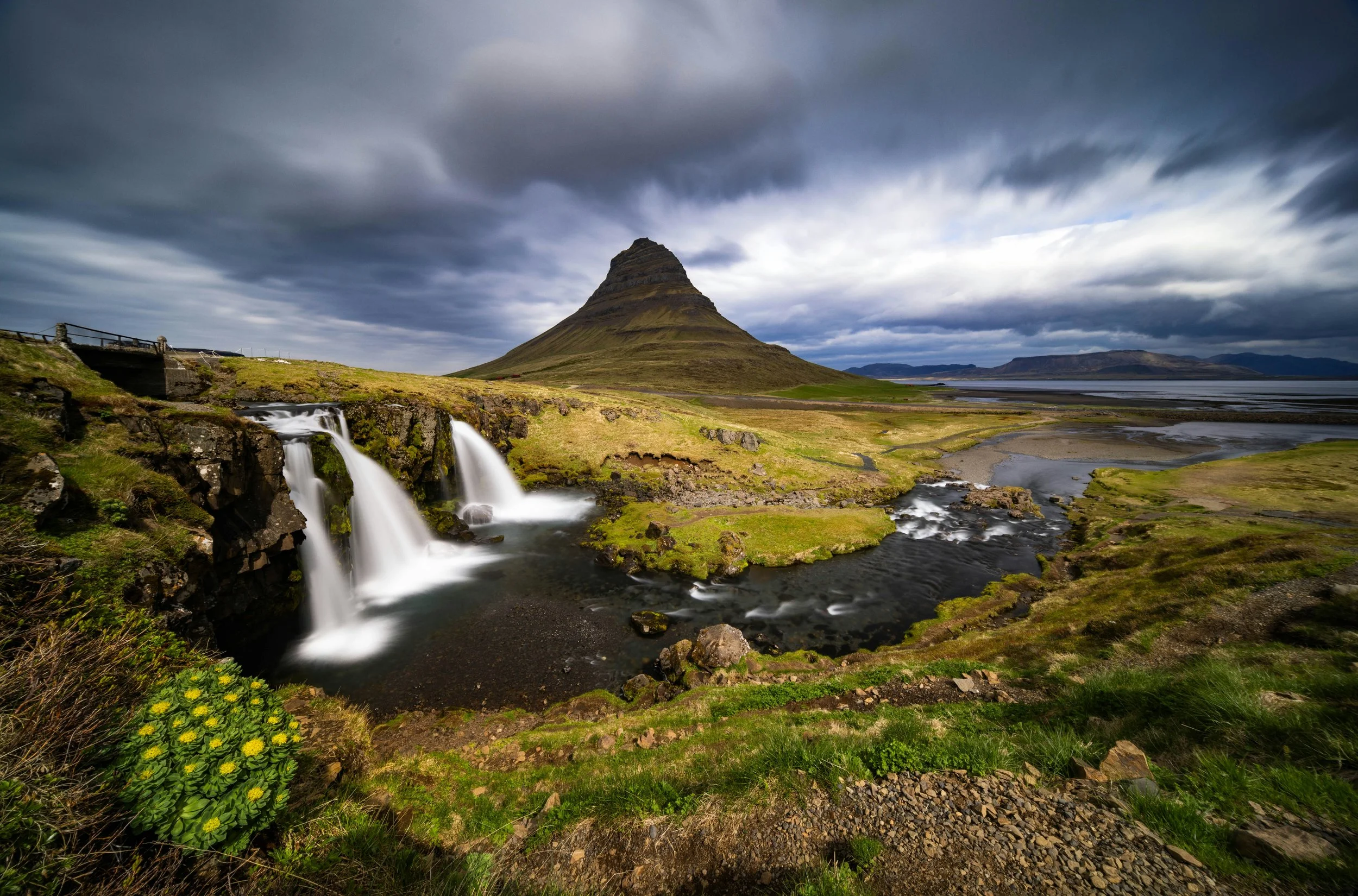 A scenic landscape featuring a tall, pointed mountain, a waterfall cascading over rocks, green grass, and cloudy skies.
