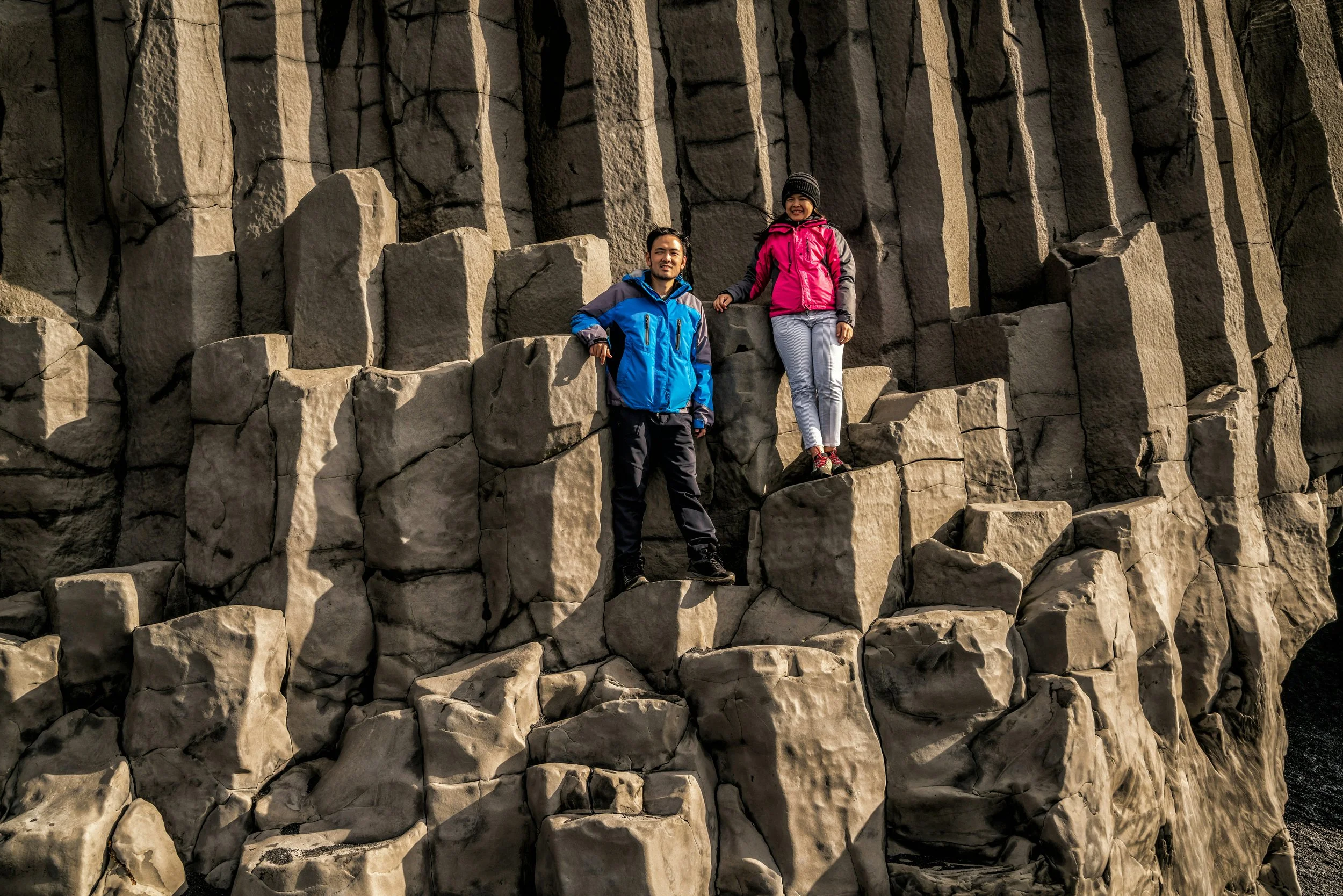 A man and a woman standing on rocks in front of tall basalt columns, smiling. The woman is wearing a pink jacket, white pants, and a beanie, while the man is wearing a blue and black jacket and dark pants.