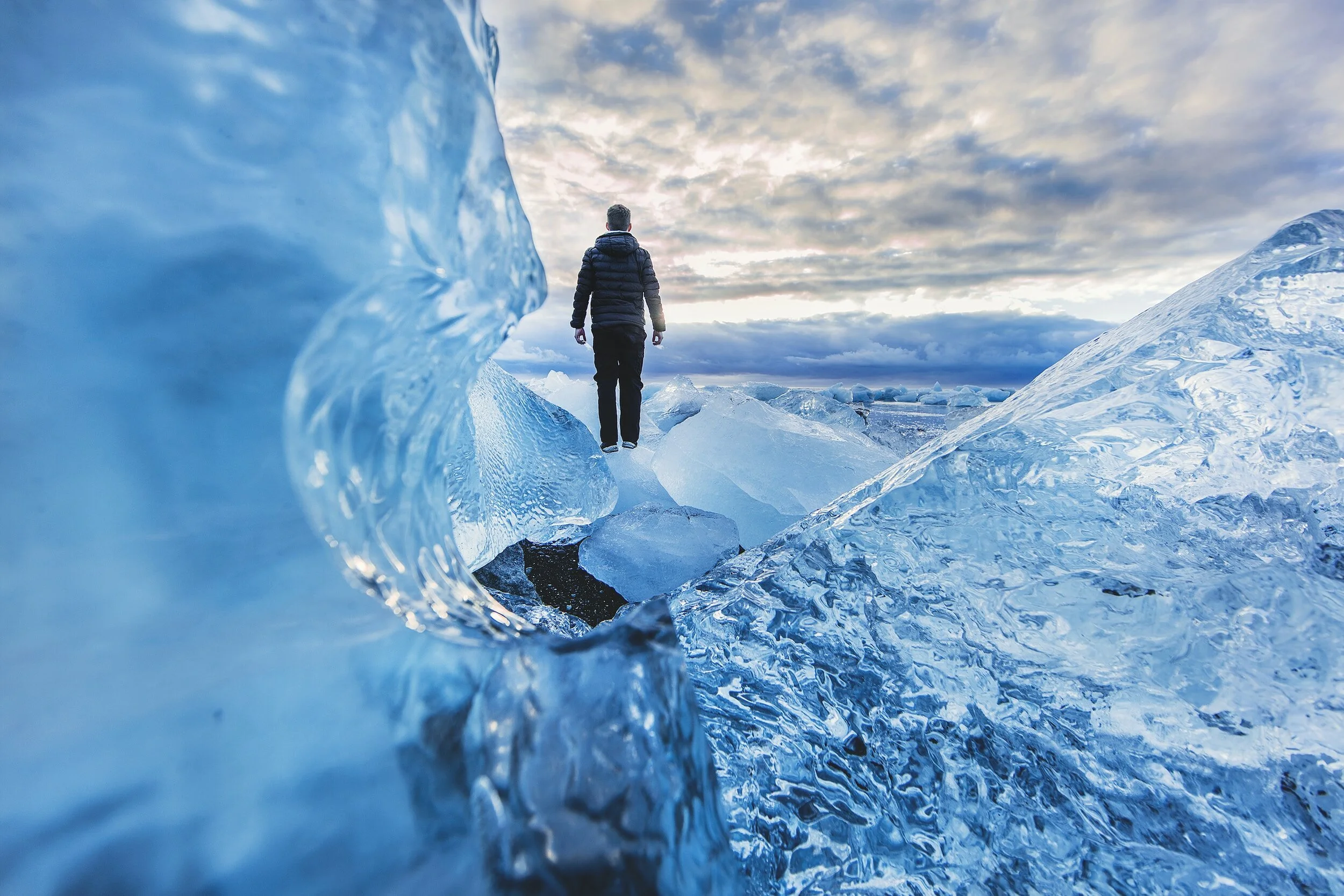 Person standing on icy terrain surrounded by large ice formations, under a cloudy sky.