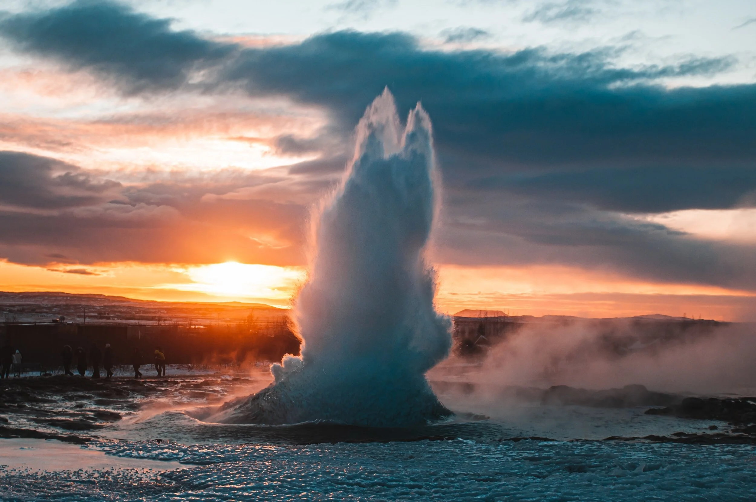 Geyser erupting with sunset in background, steam rising, silhouetted people watching.