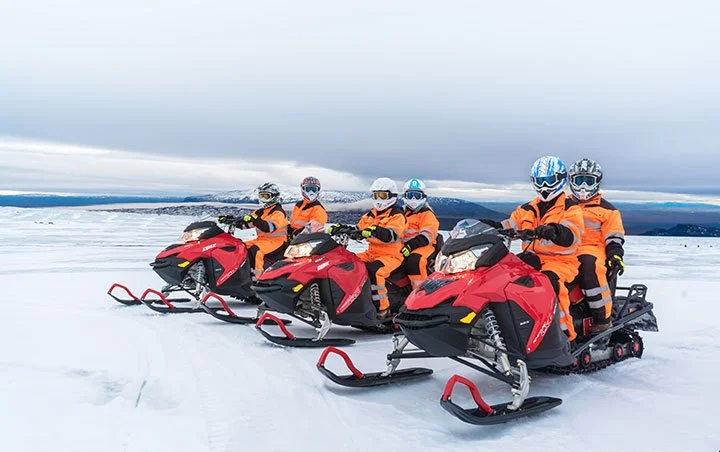 Group of people wearing orange jackets riding red snowmobiles on a snowy landscape.