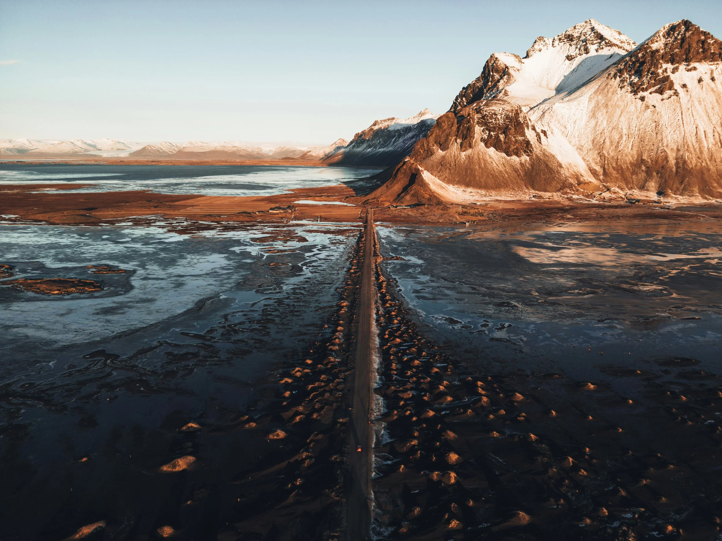 A mountainous landscape featuring snow-capped peaks, a river or lake at the foot, with a dirt road leading towards the mountains, under a clear sky.