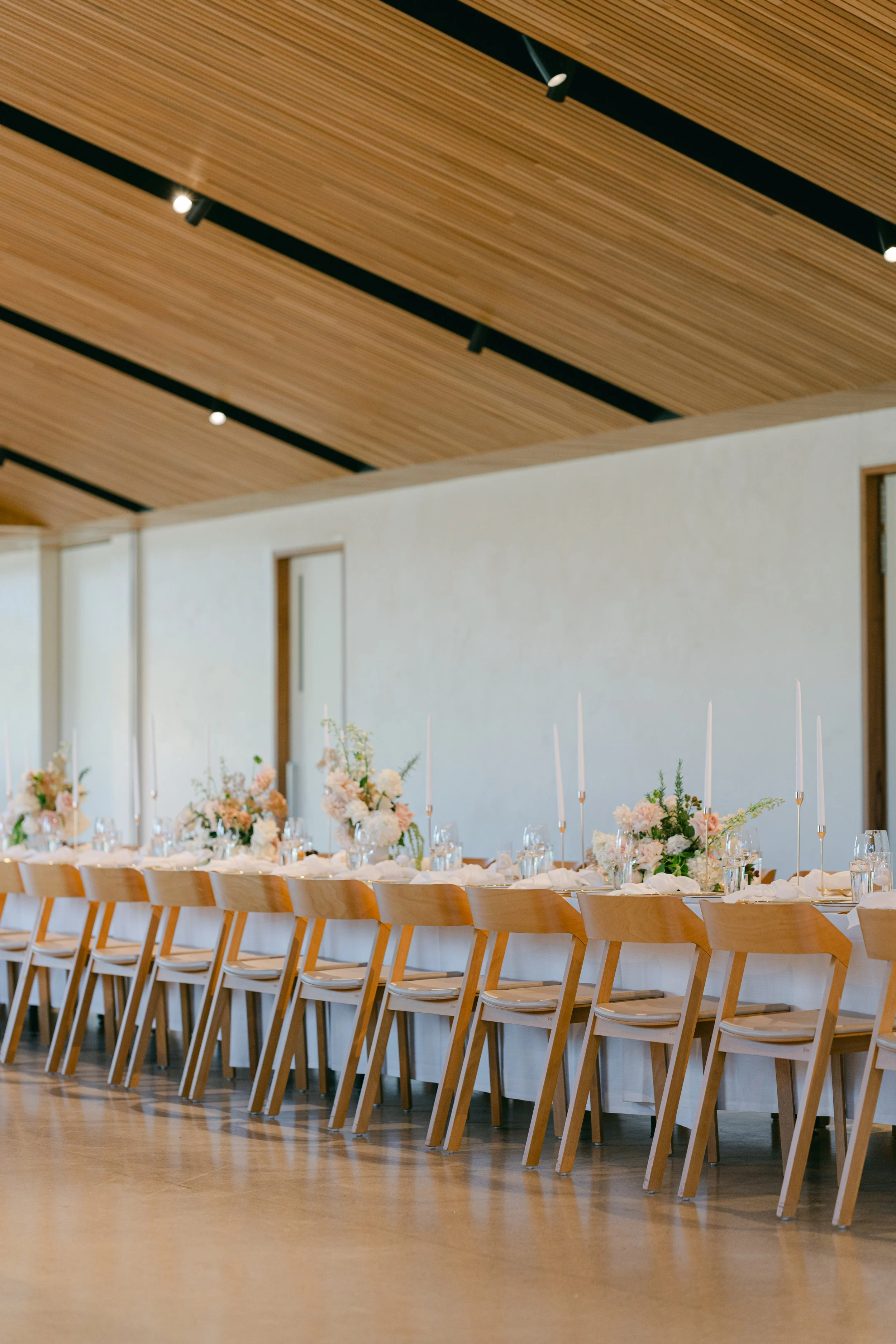Elegant banquet table decorated with pastel floral arrangements and tall white candles in a modern room with wooden ceiling.