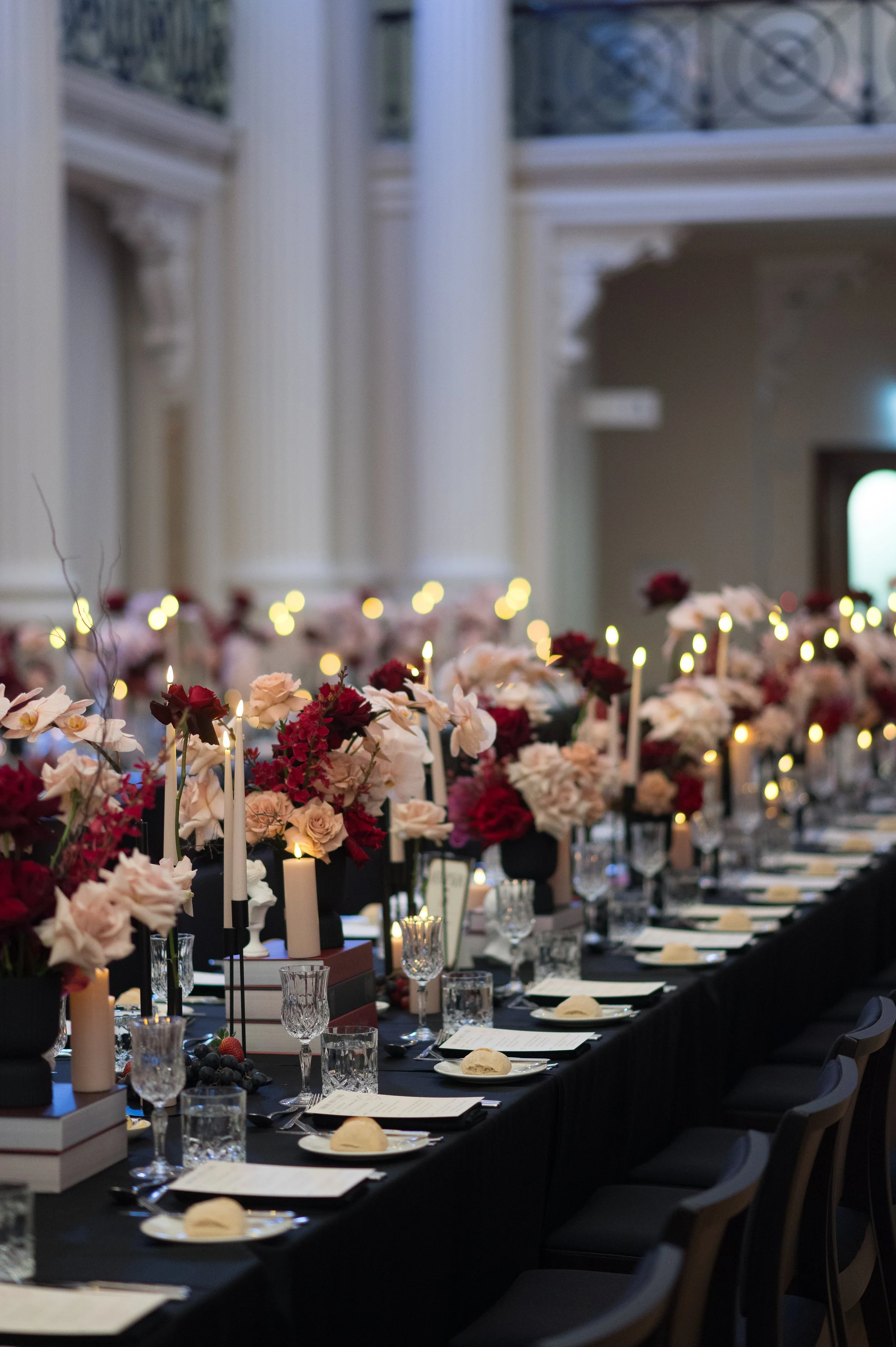 Elegant banquet table set with floral arrangements, candles, and tableware in a grand hall with white columns and high ceilings.