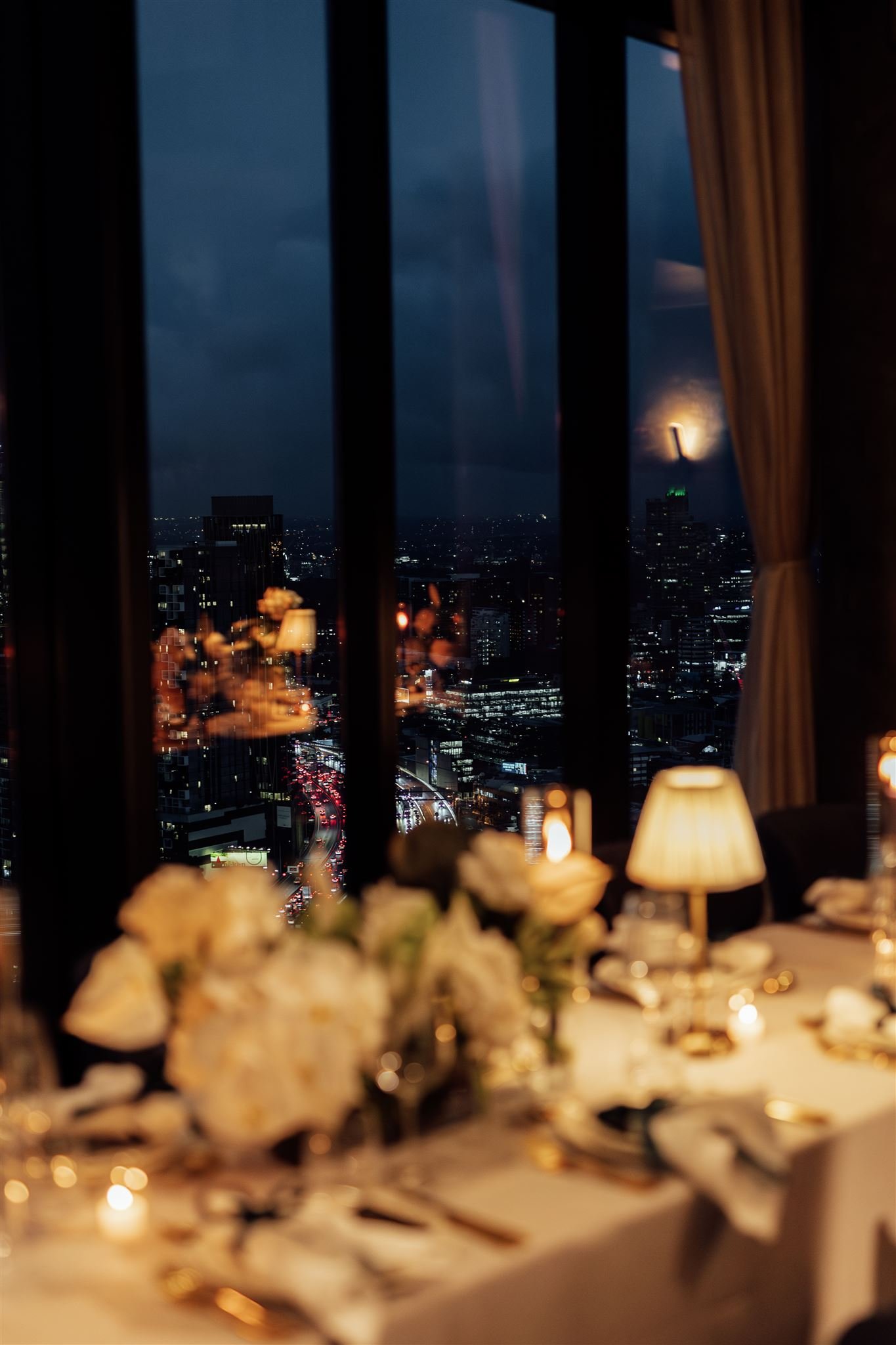 Nighttime cityscape view through a window from a decorated dining table with flowers, lamps, and dinnerware.