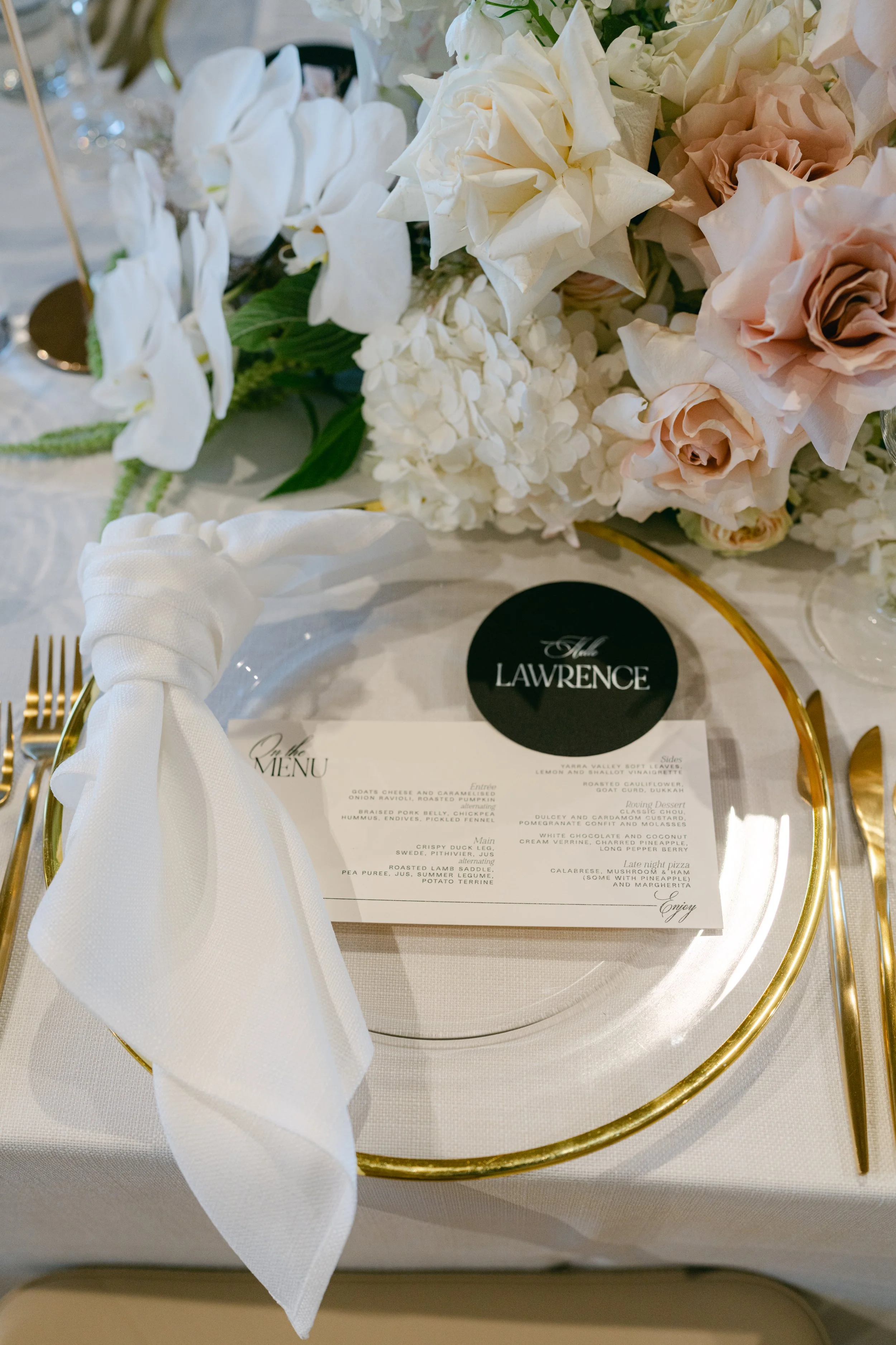 Elegant table setting with a gold-rimmed glass charger, white napkin, and a floral centerpiece featuring white and blush pink flowers. There is a black and white menu card with the name 'Lawrence' at the top, placed on a clear glass plate. Gold flatw