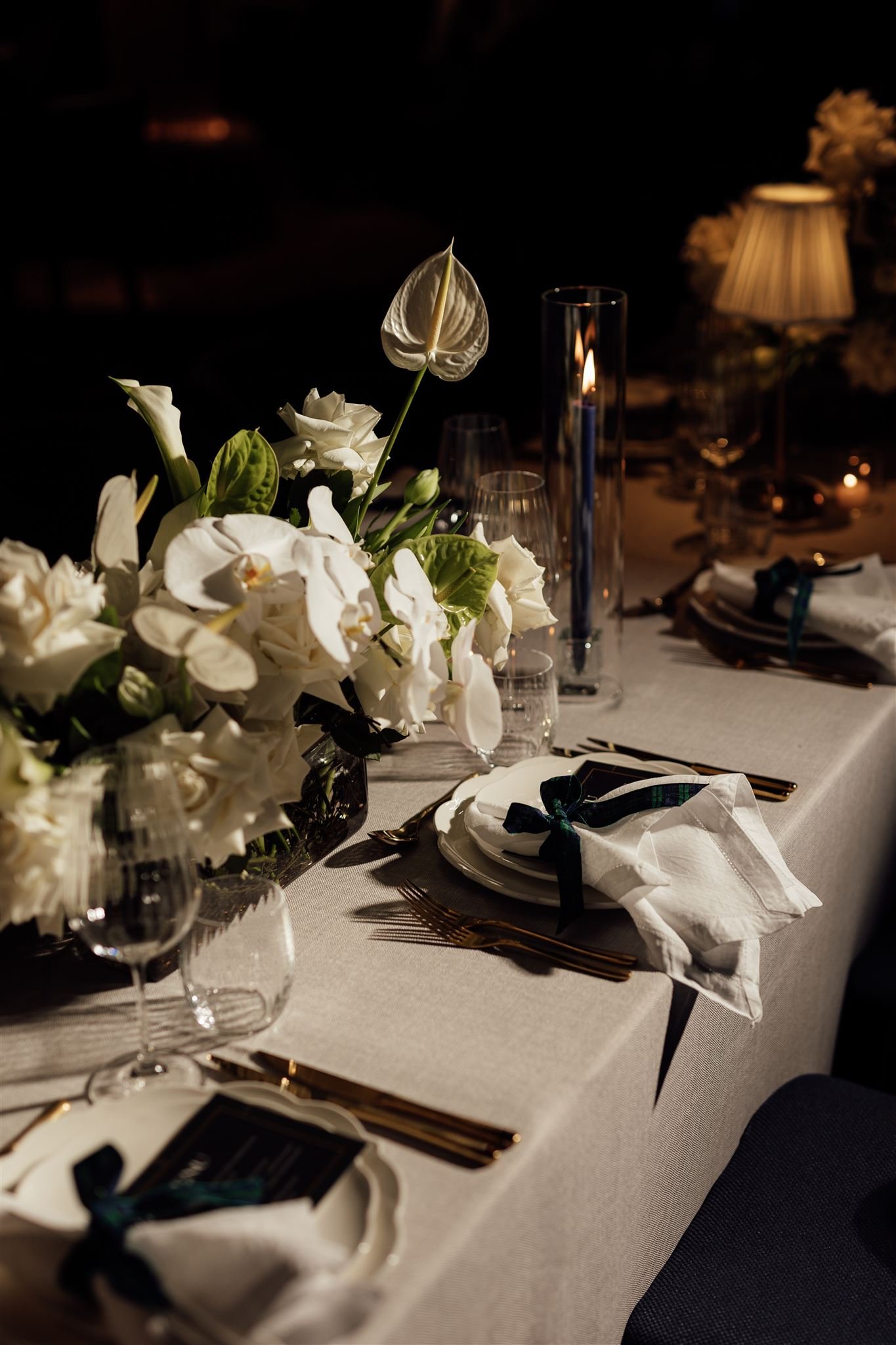 Elegant dining table setup with white flowers, candles, wine glasses, and gold utensils in a dimly lit room.