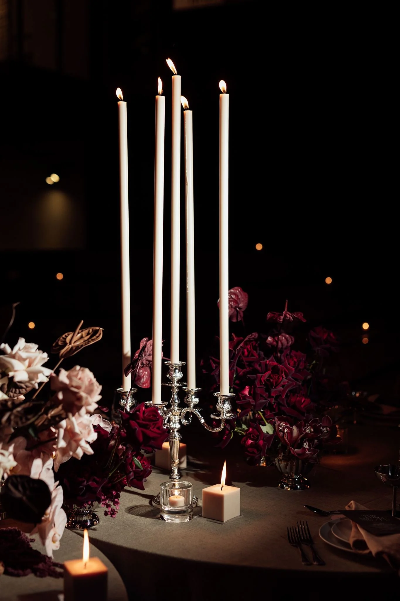 A dark dining table setting with tall white candles in silver candle holders, surrounded by pink and red flowers, with some lit candles on the table.