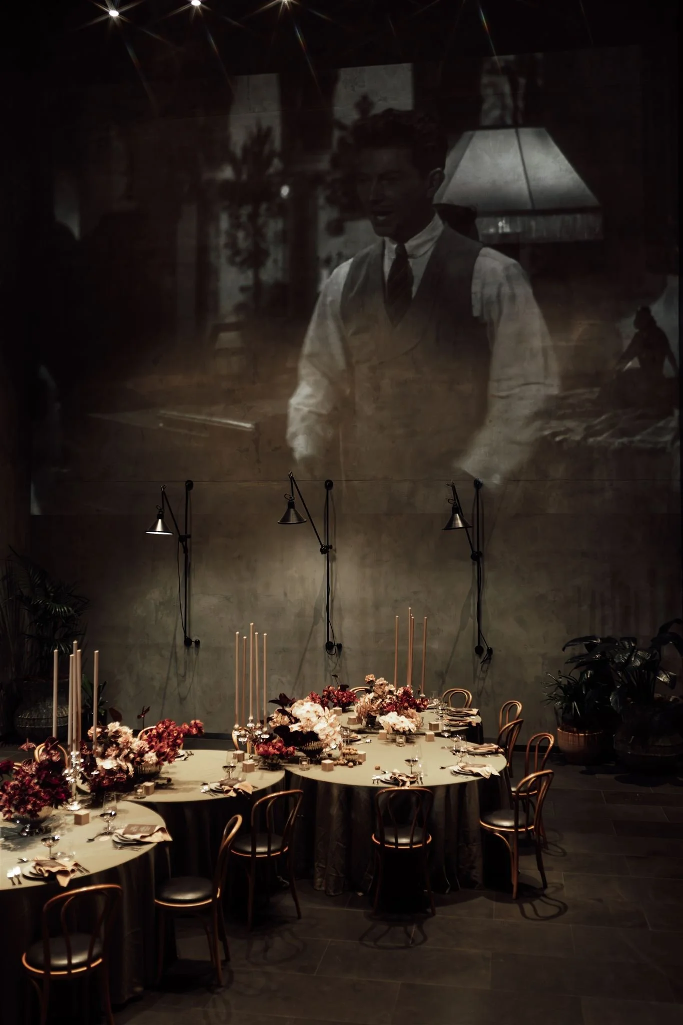 Elegant dining setup with multiple round tables adorned with flowers, candles, and place settings, in a dimly lit room with a large black-and-white film projected on the wall showing a man in formal attire.