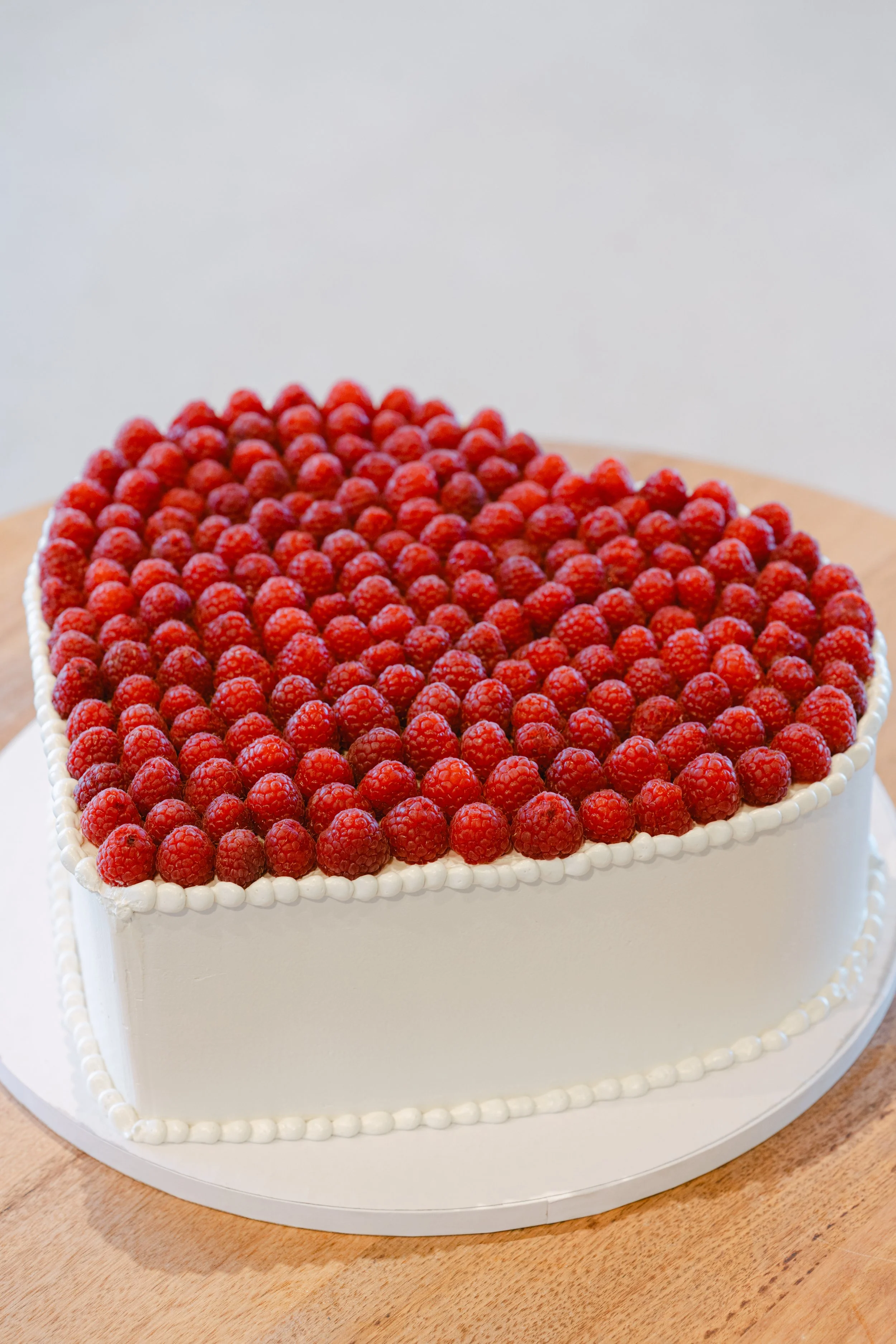 Heart-shaped cake topped with fresh raspberries and decorated with white piping, sitting on a wooden surface.