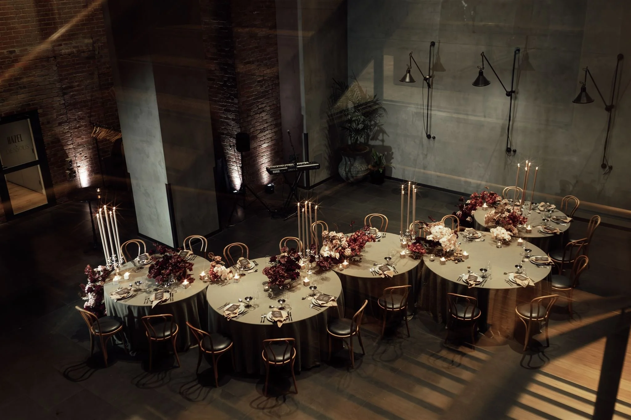 Elegant event space with three round tables decorated with floral centerpieces, candles, and place settings, set for a formal dinner. The space features exposed brick and concrete walls, wall-mounted lamps, and a small stage with a keyboard and micro
