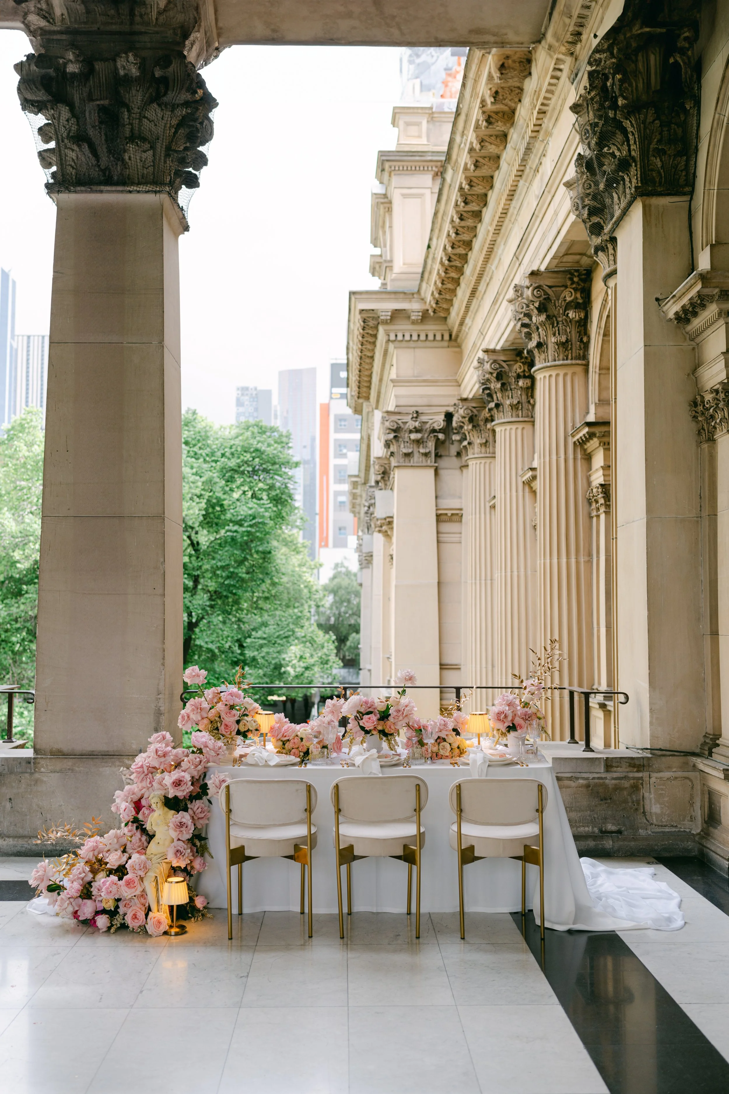 Elegant wedding reception table with pink flowers and candles on a balcony with classical architecture and city skyline in the background.