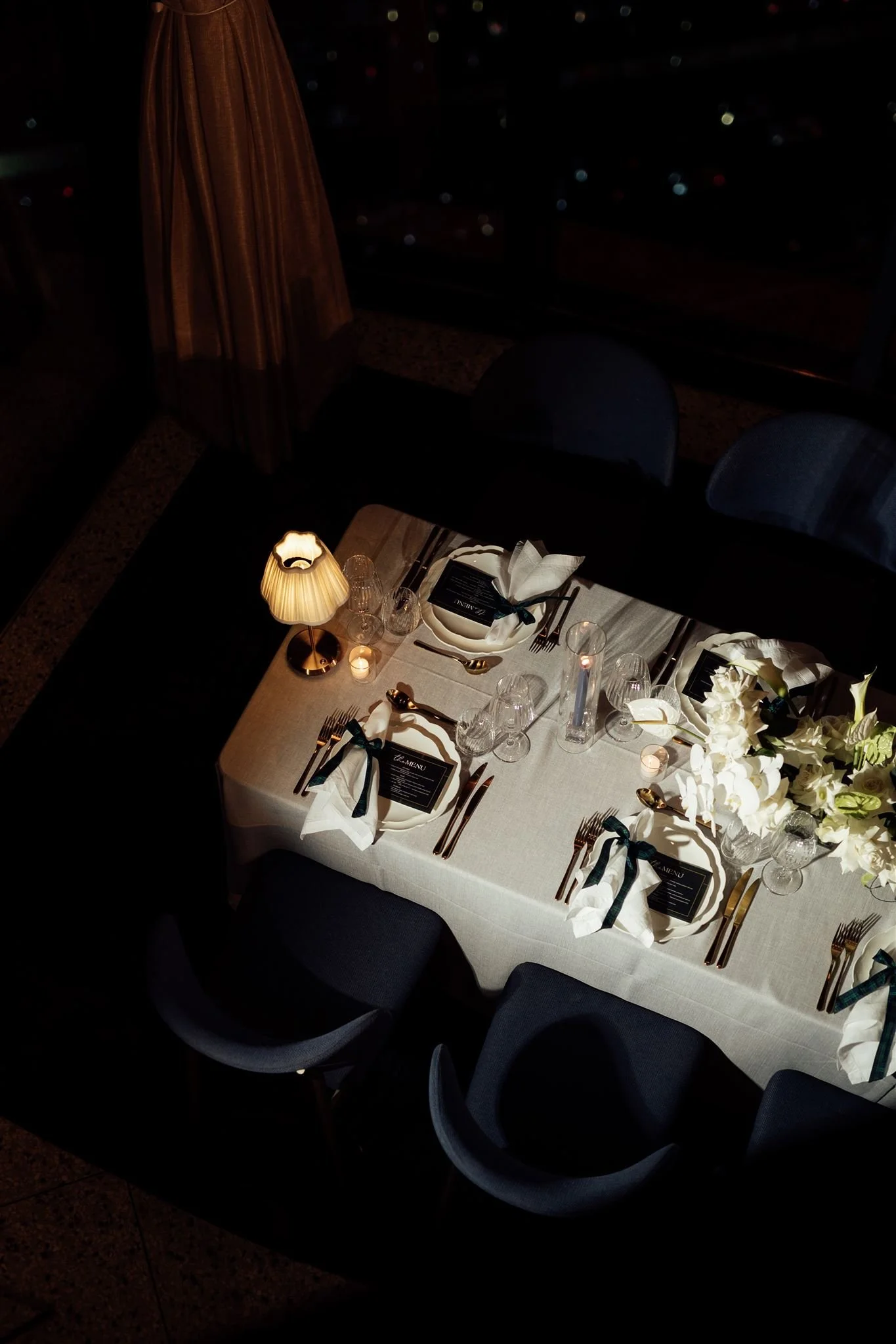 Elegant dining table set for a formal event, with white tablecloth, arranged dinnerware, glasses, black and white napkins, a floral centerpiece, candles, and a table card indicating it is a dinner menu.