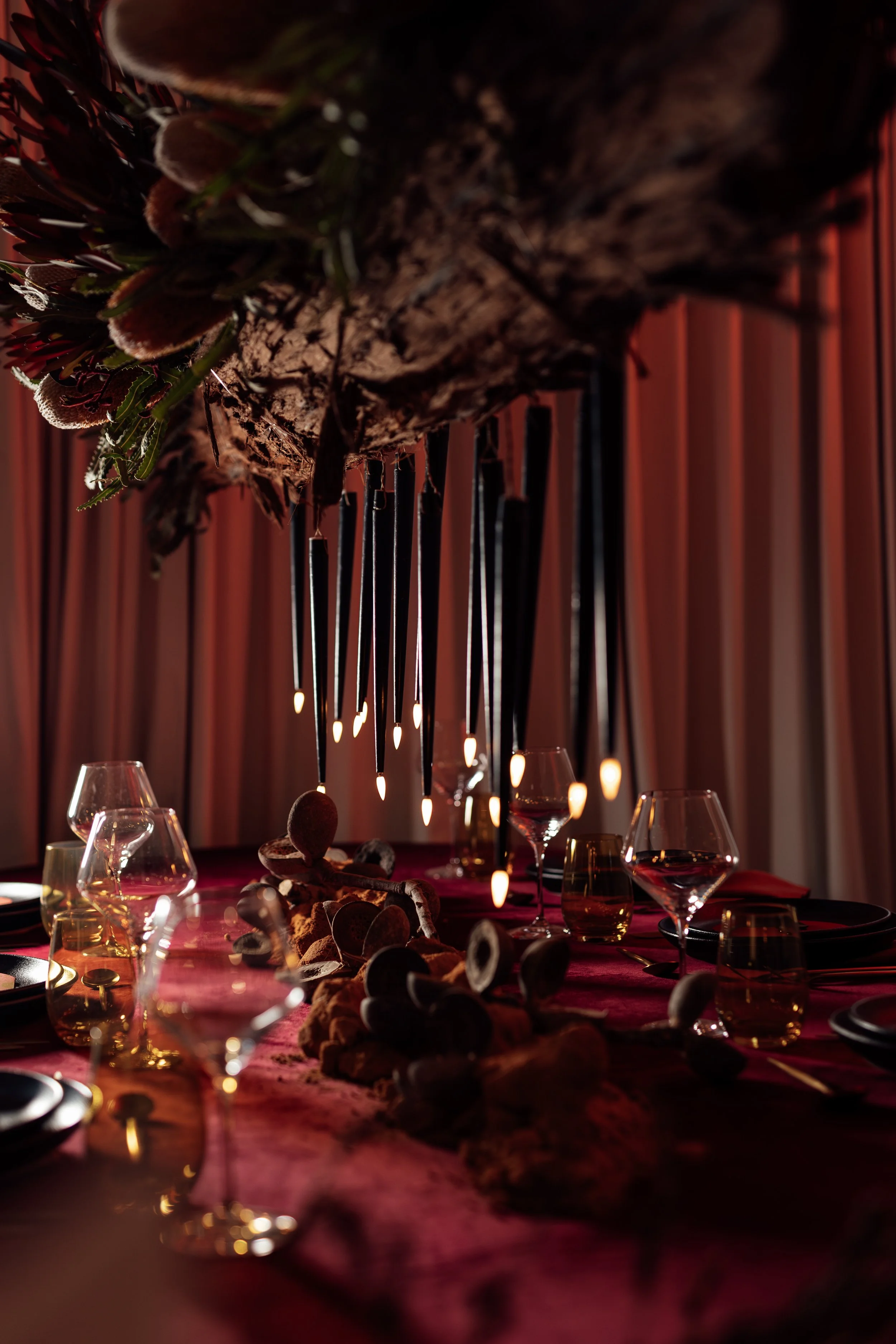 A dimly lit banquet table decorated with a dark red tablecloth, wine glasses, and candles, with a large wooden beam overhead adorned with greenery and candles hanging. The background features long curtains, creating an elegant and cozy atmosphere.