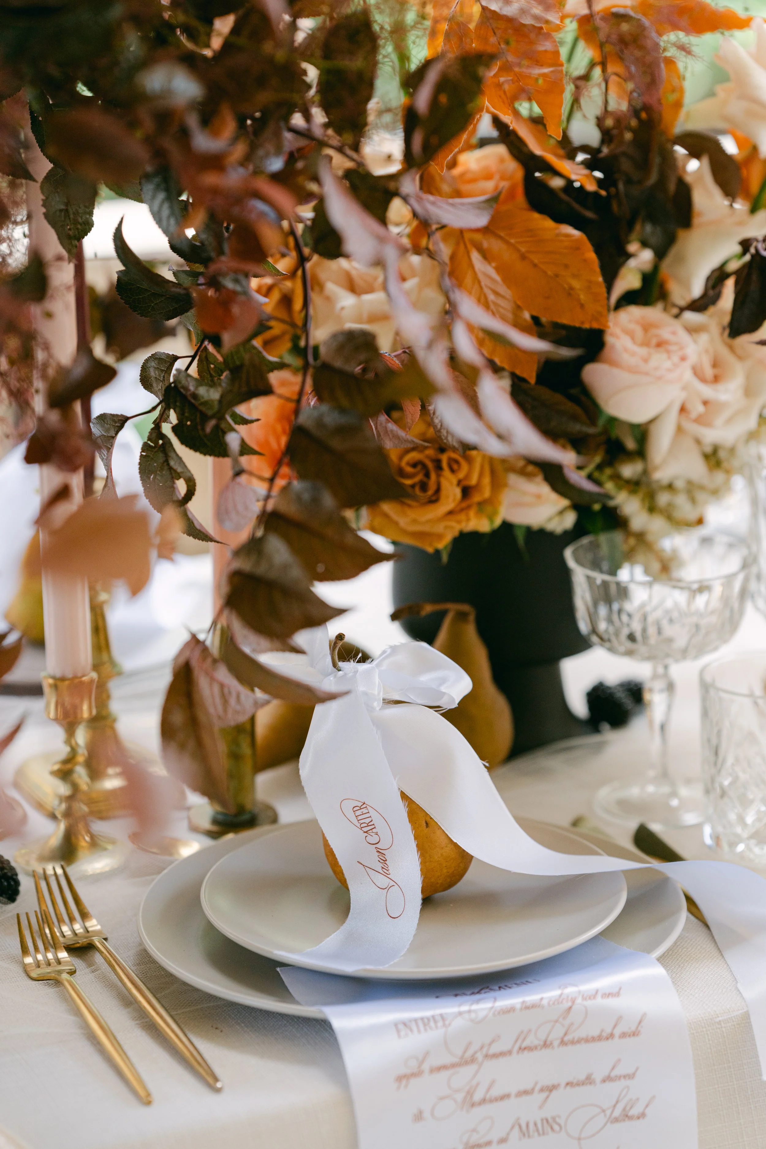 A wedding tablescape with a floral centerpiece featuring orange and white flowers, leafy accents, and a black vase. There are gold utensils, a white plate with an orange dessert tied with a white ribbon that has a name written on it, and a printed me