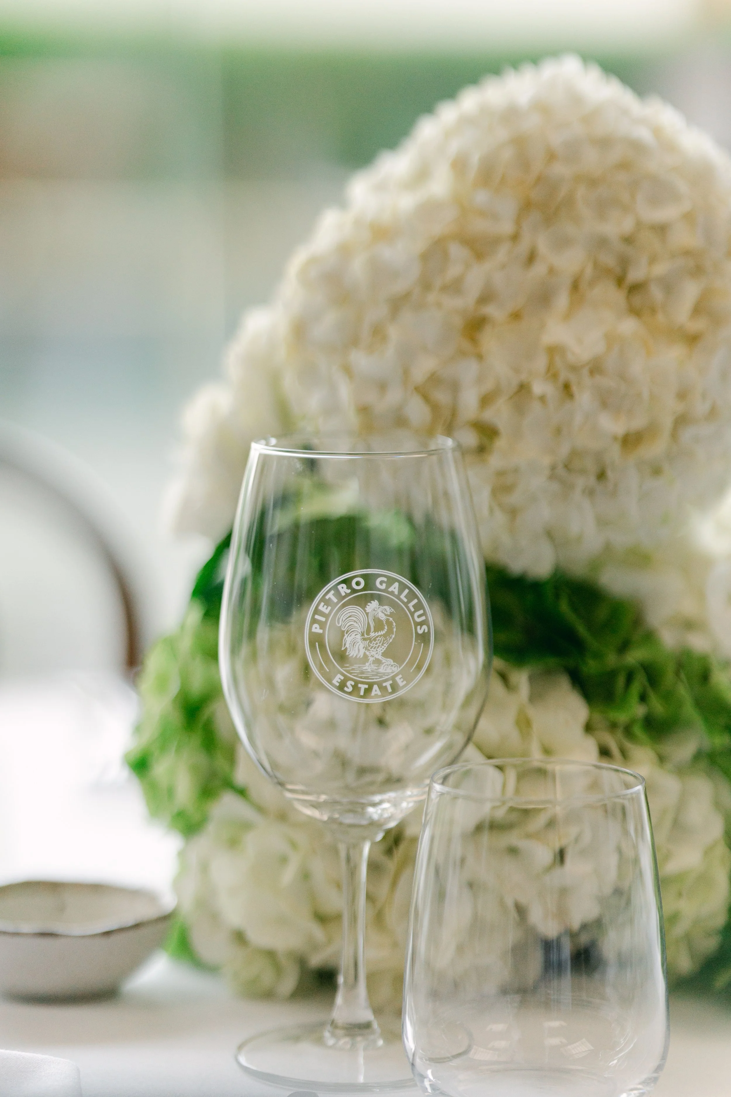 A clear wine glass with the logo 'Pietro Gallicus Estate' engraved on it, placed in front of a large bouquet of white flowers on a table.