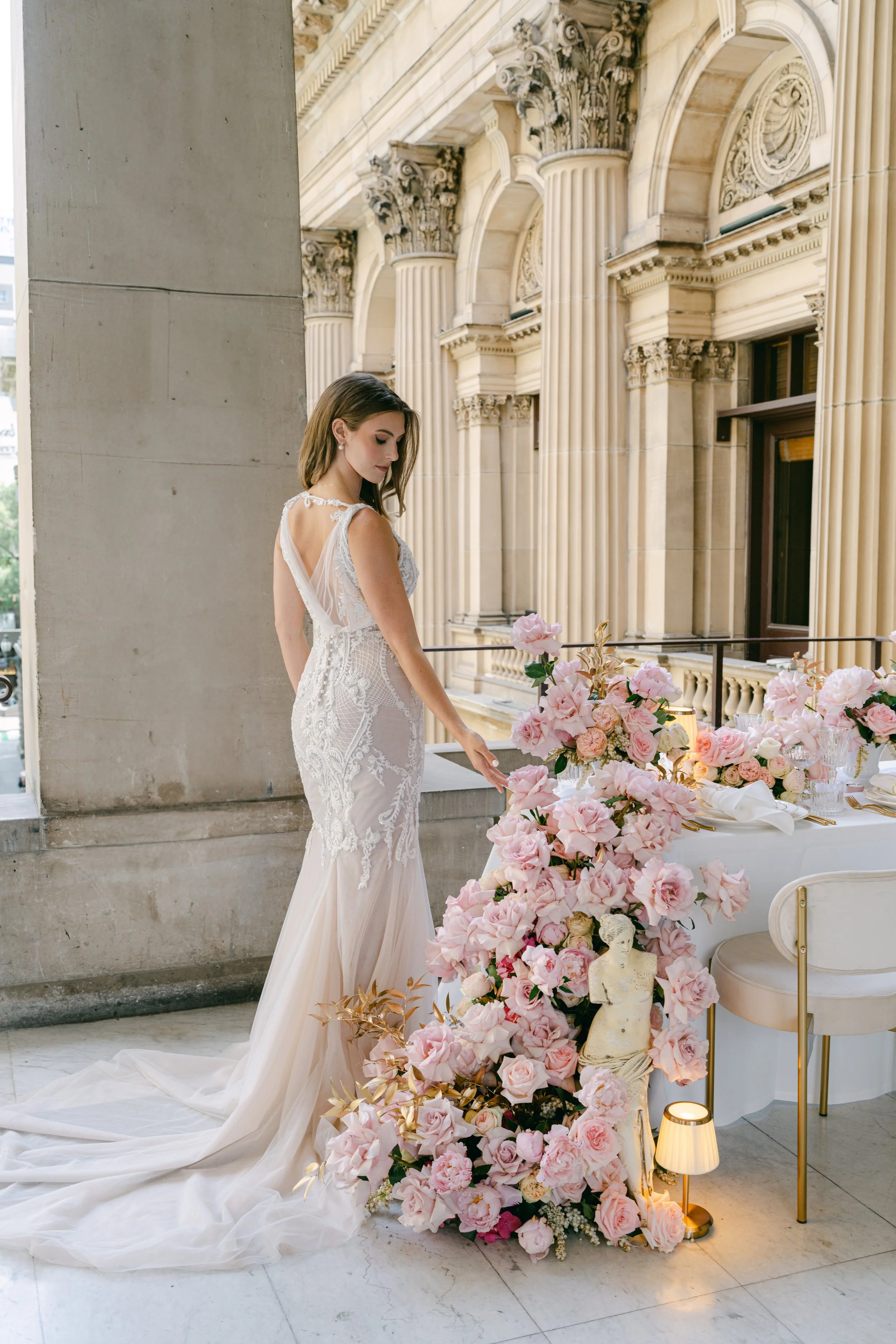 A woman in a white lace wedding dress stands beside a table decorated with pink roses and a small classical statue, in a grand neoclassical building with tall columns.