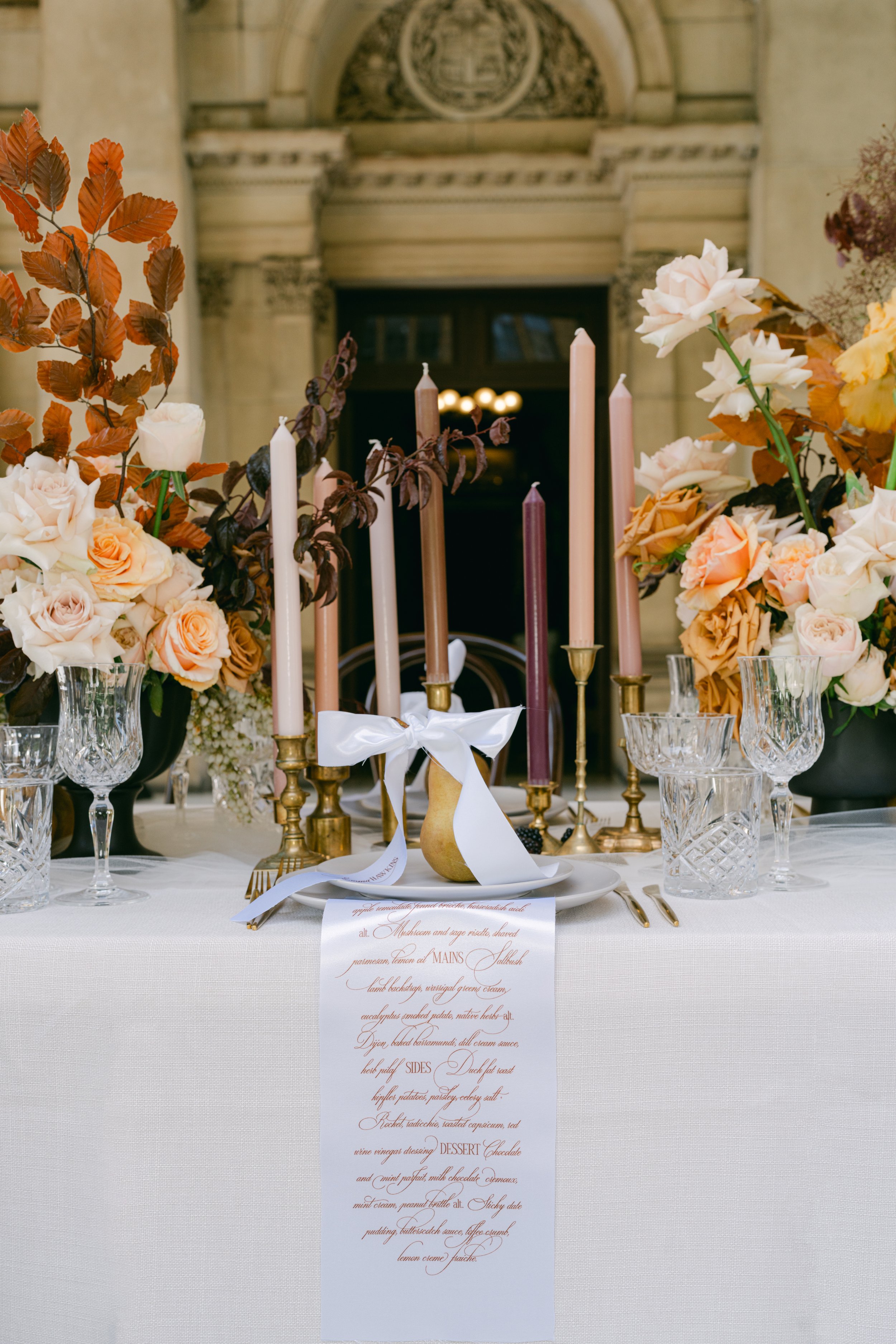 Elegant table setting with pink and white roses, candles, glassware, and a menu card in a formal event space.