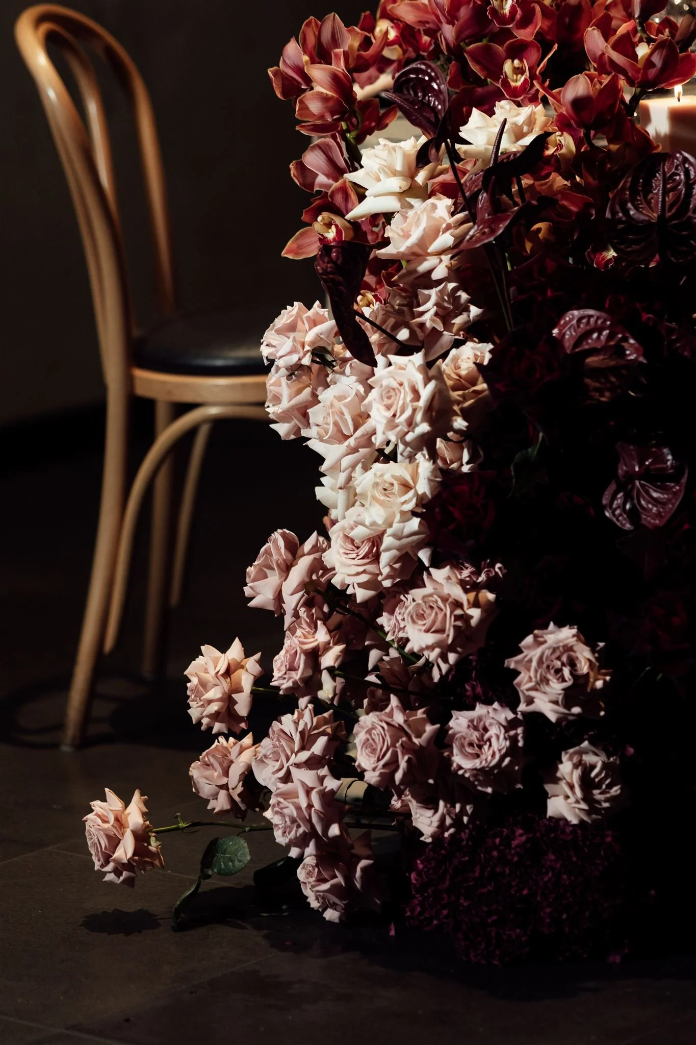 A large arrangement of pink and white roses and dark purple and burgundy flowers on a dark floor, with a beige chair in the background.