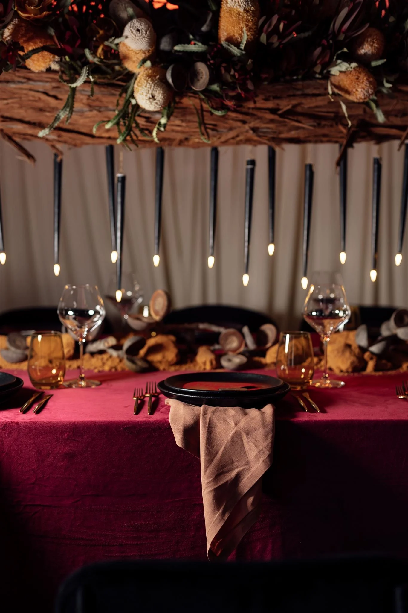 Elegant dining table set for a formal event, with black plates, gold utensils, wine glasses, amber-colored glasses, a burgundy tablecloth, and a beige napkin, with a rustic floral and candle centerpiece above.