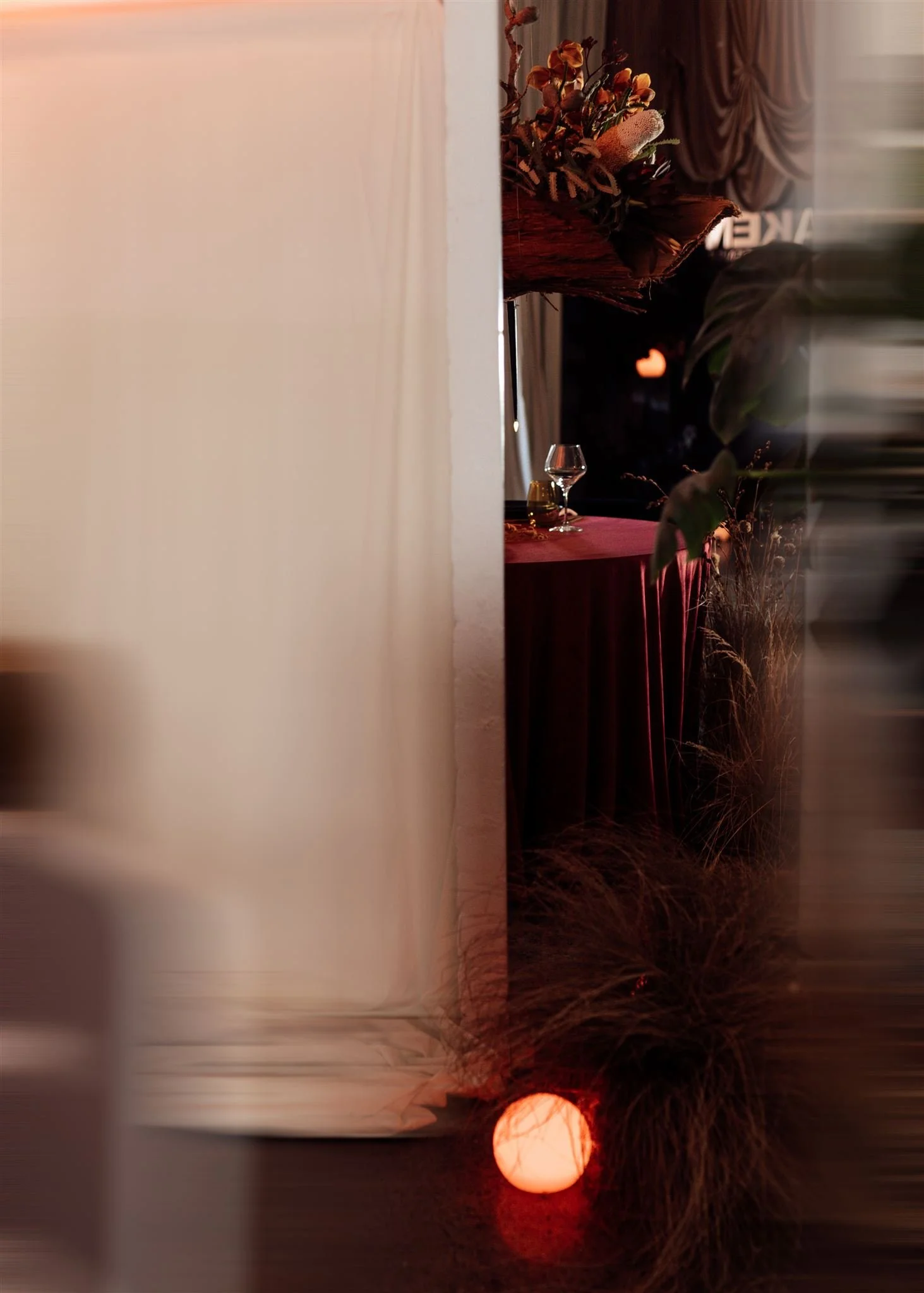 Partially hidden view of a decorated indoor event space with a table draped in red cloth, wine glasses, floral arrangements, and warm lighting, seen through a partially opened white curtain.