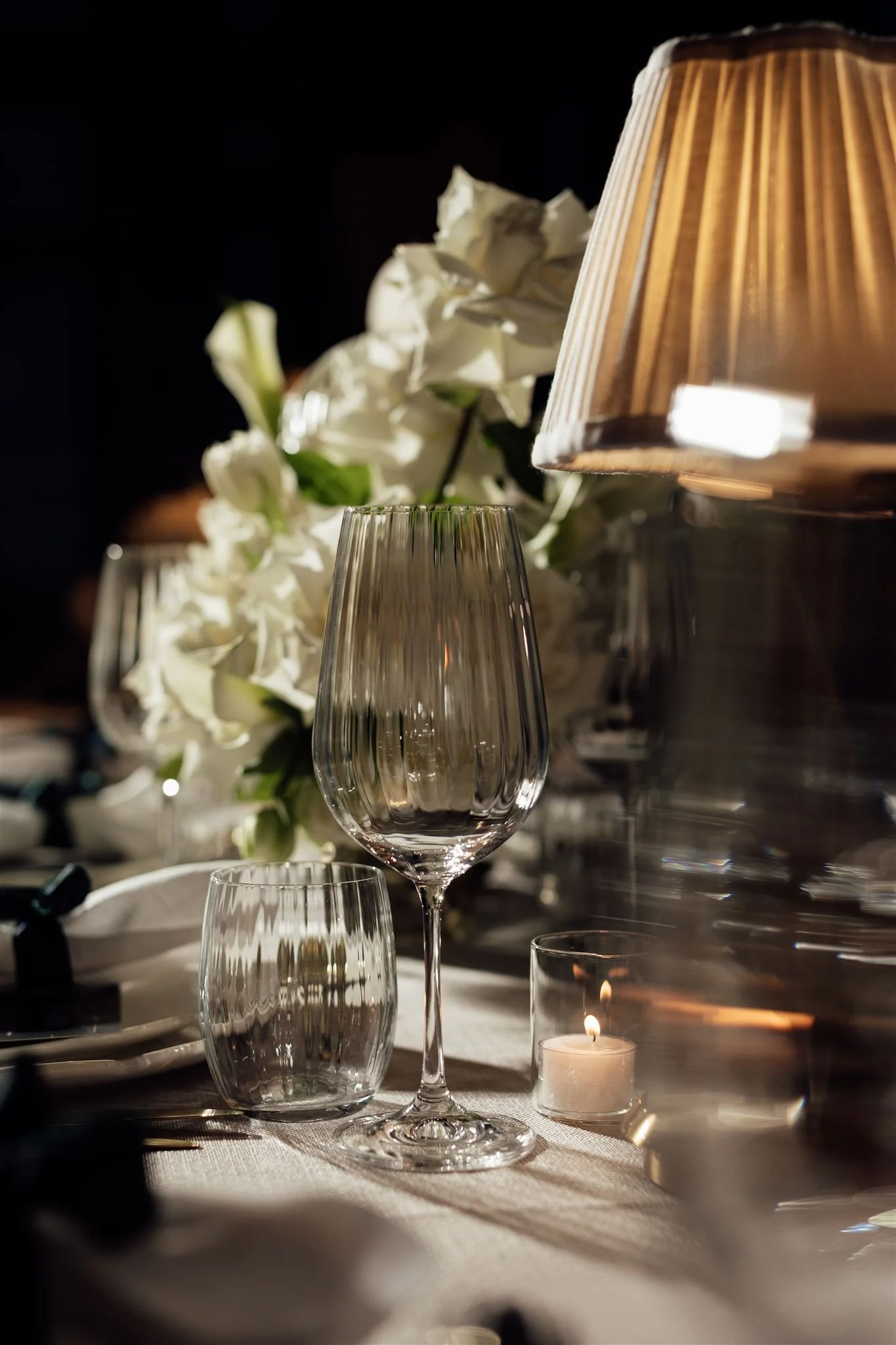 Elegant table setting with empty wine and water glasses, a lit candle, white flowers, and a lamp on a white tablecloth.