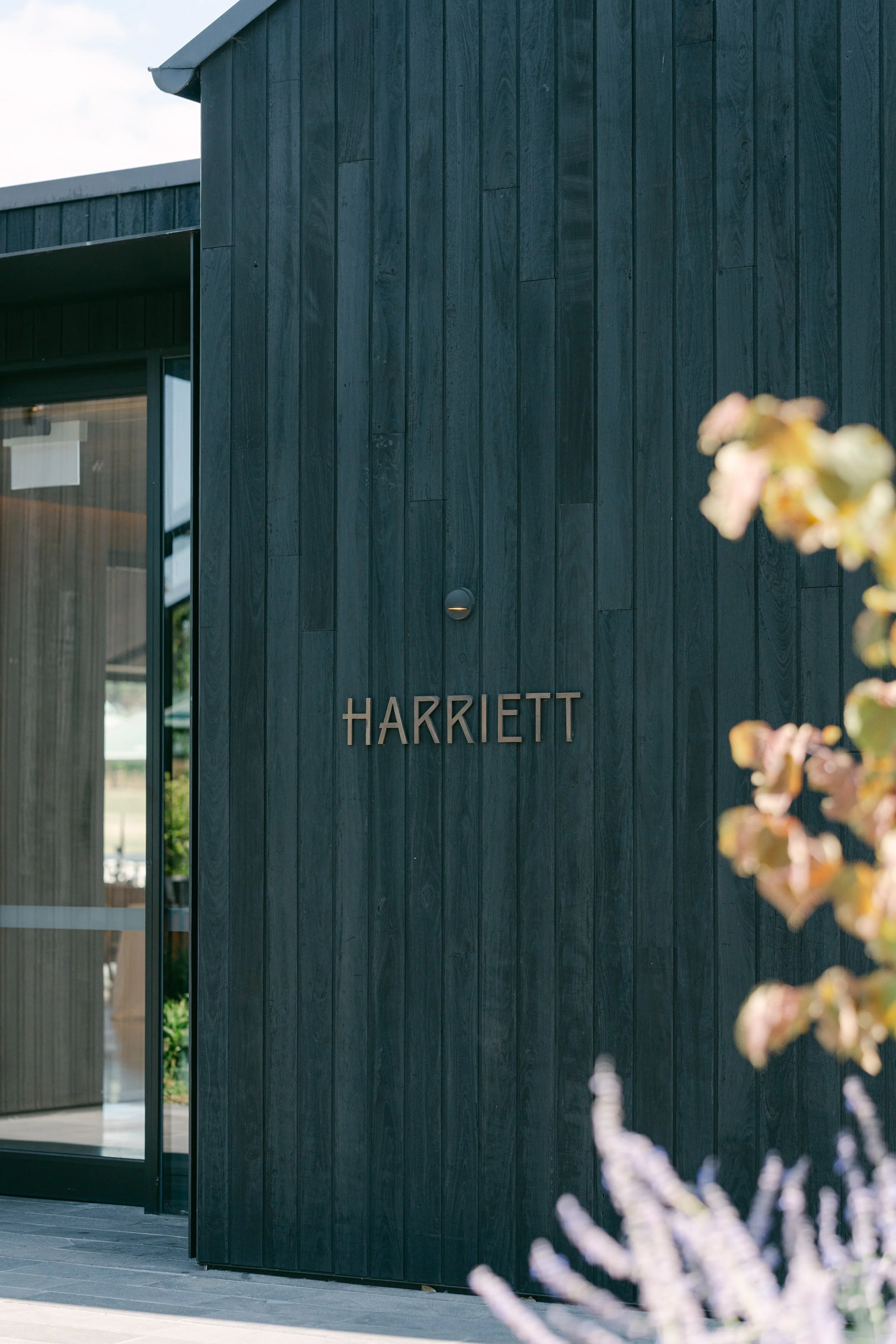 Exterior of a building with black wooden siding and the name 'HARRIETT' on the wall next to a glass door.