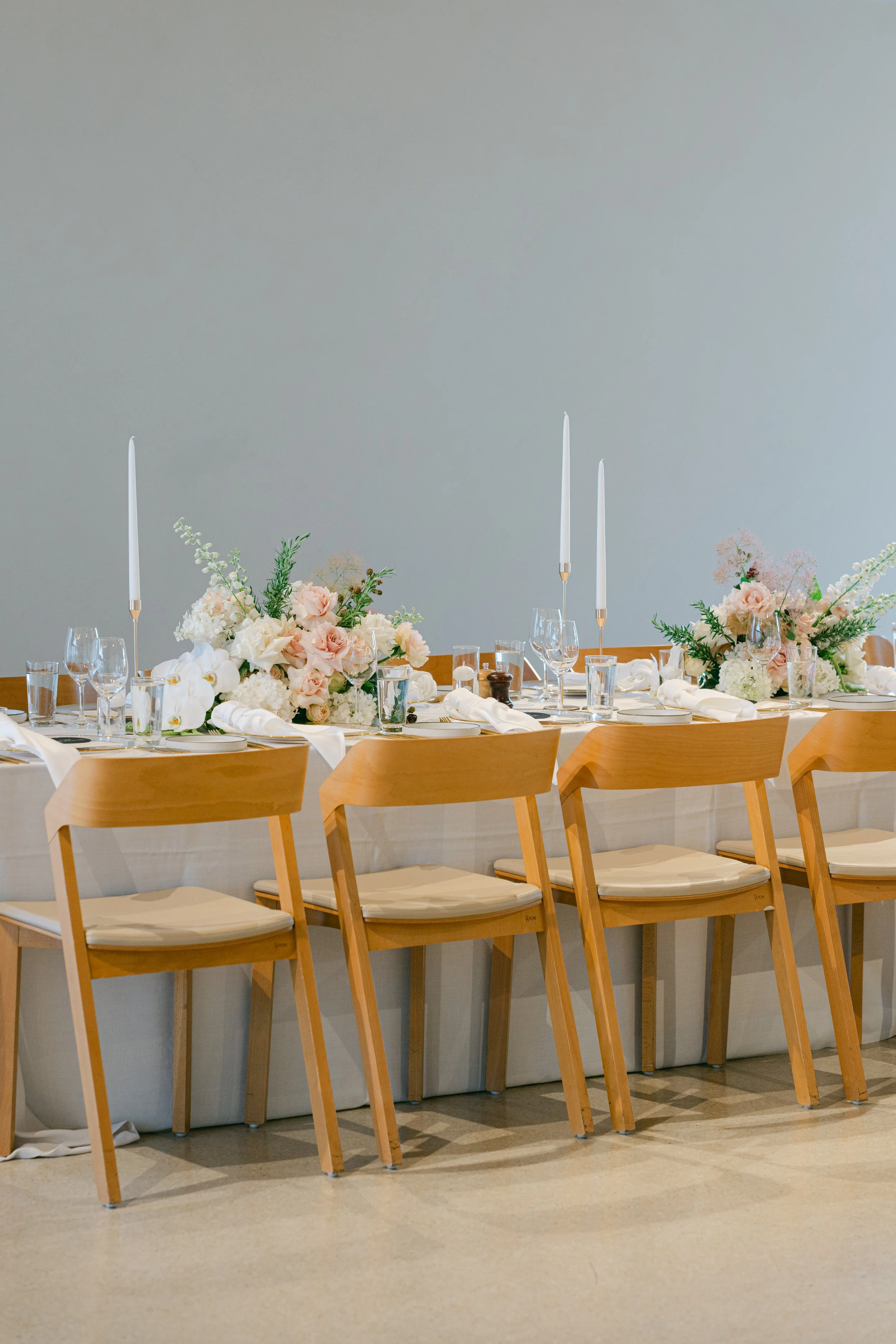 Elegantly set dinner table with floral centerpieces, candles, glassware, and napkins, arranged on a long table in a neutral-colored room.