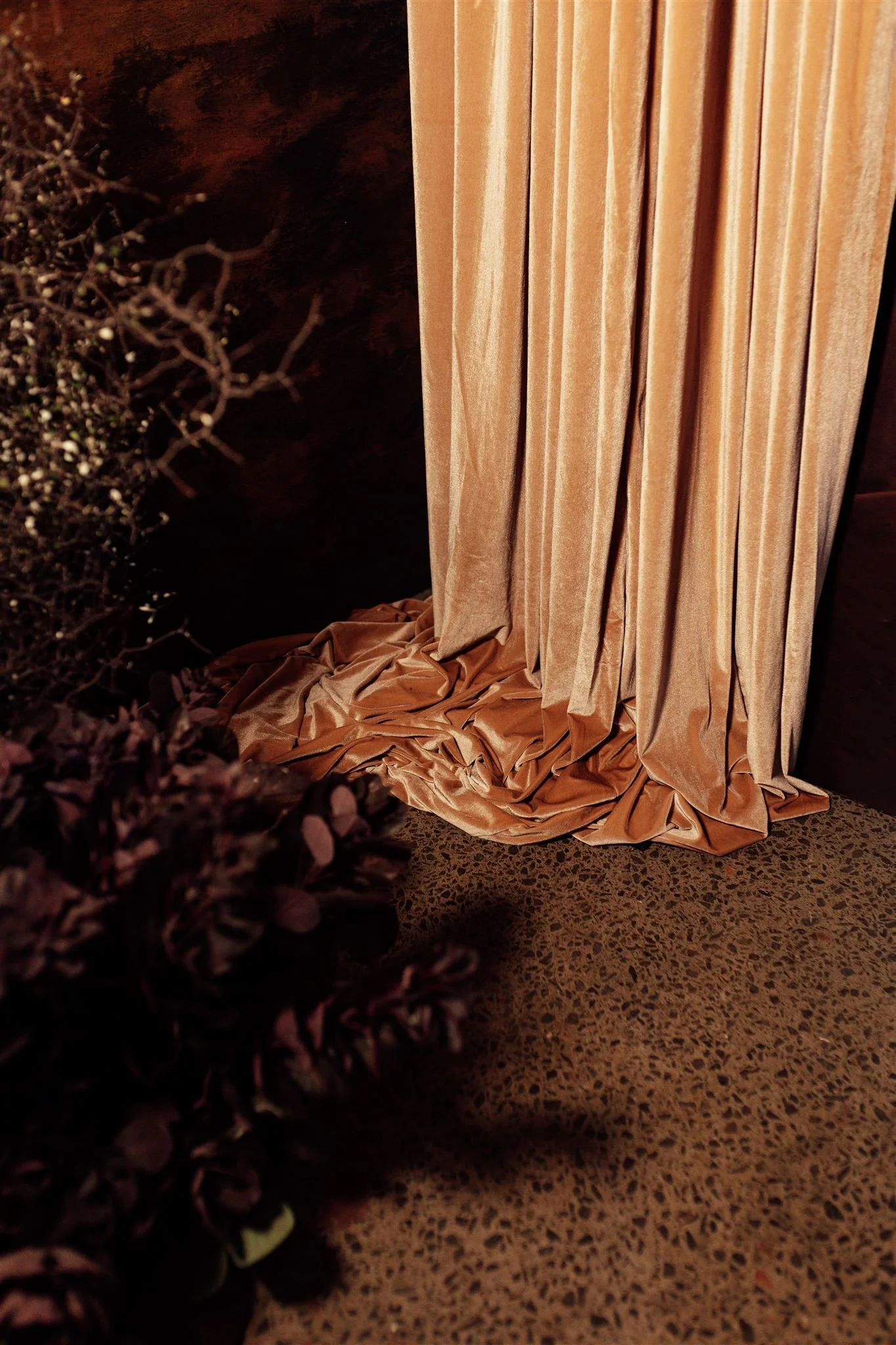 A beige velvet curtain partially pulled aside, revealing a dark wall and a floral arrangement with purple flowers on the floor.