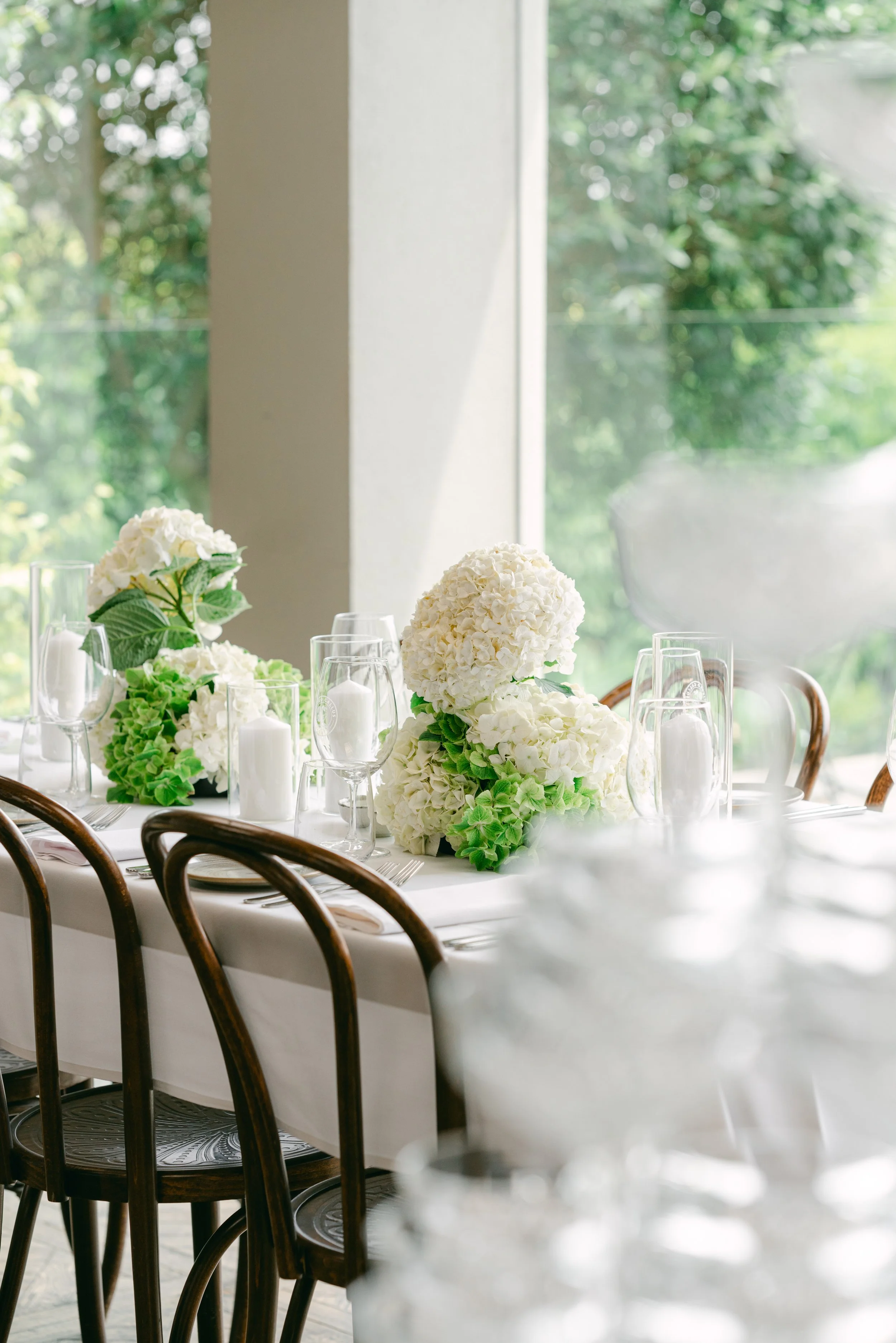 Elegant dining table decorated with white and green hydrangea flower arrangements, surrounded by wine glasses and white candles, set near a large window with greenery outside.