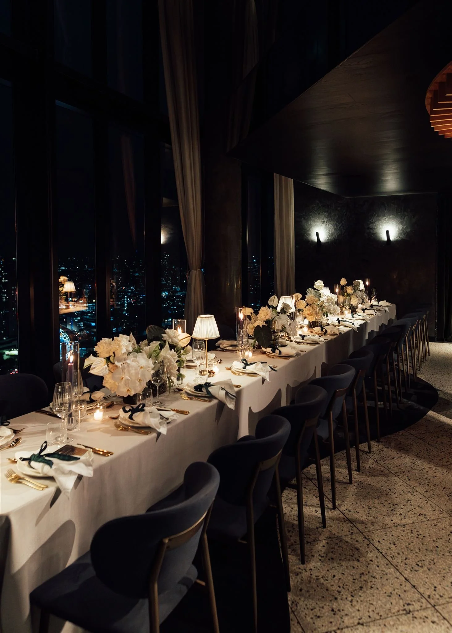 Elegant dining table set for a formal event in a tall glass-walled room at night, with city lights visible outside. The table is decorated with white flowers, candles, and tableware, and surrounded by dark chairs.