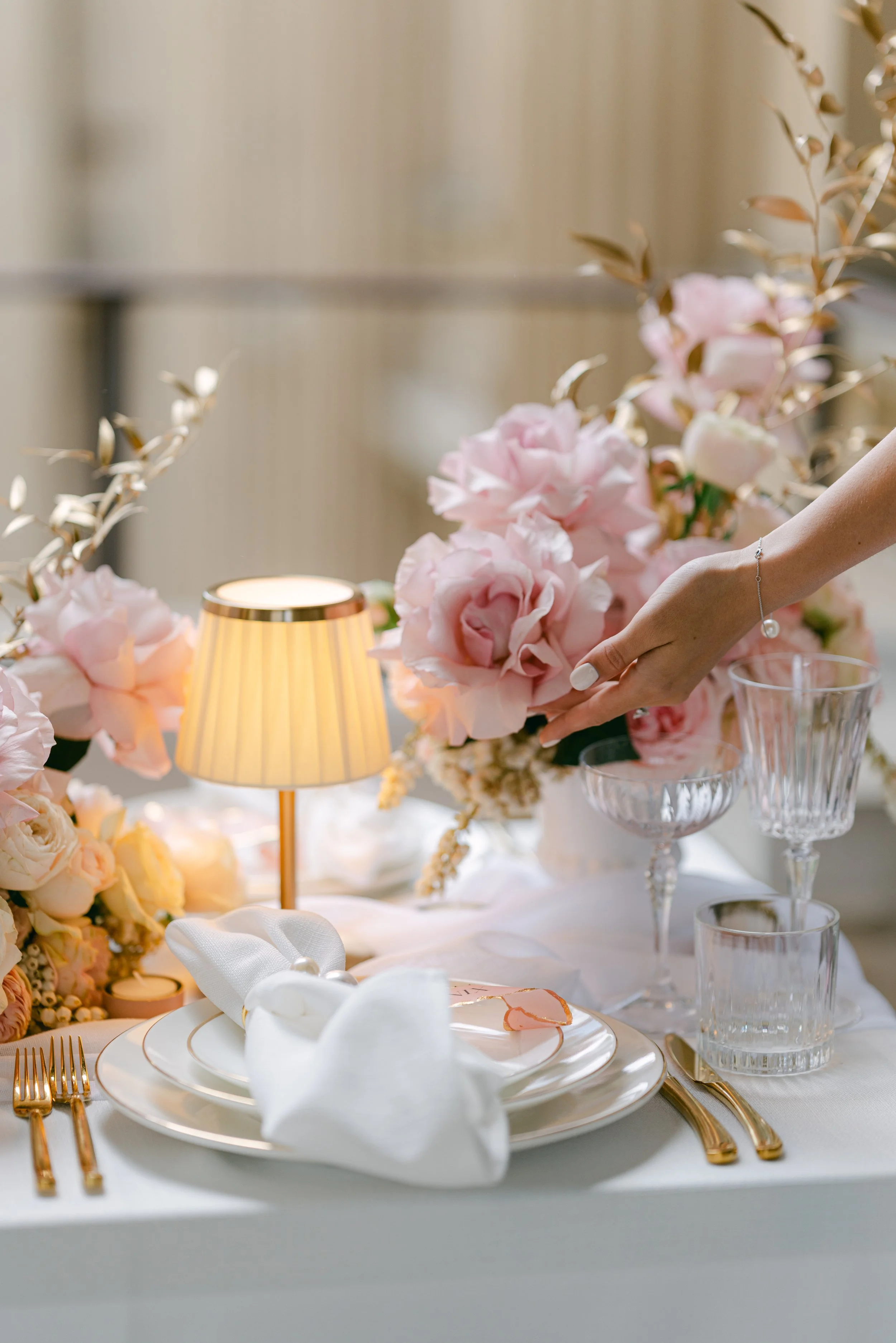 A table setting with pink flowers, glassware, gold cutlery, and a small table lamp.